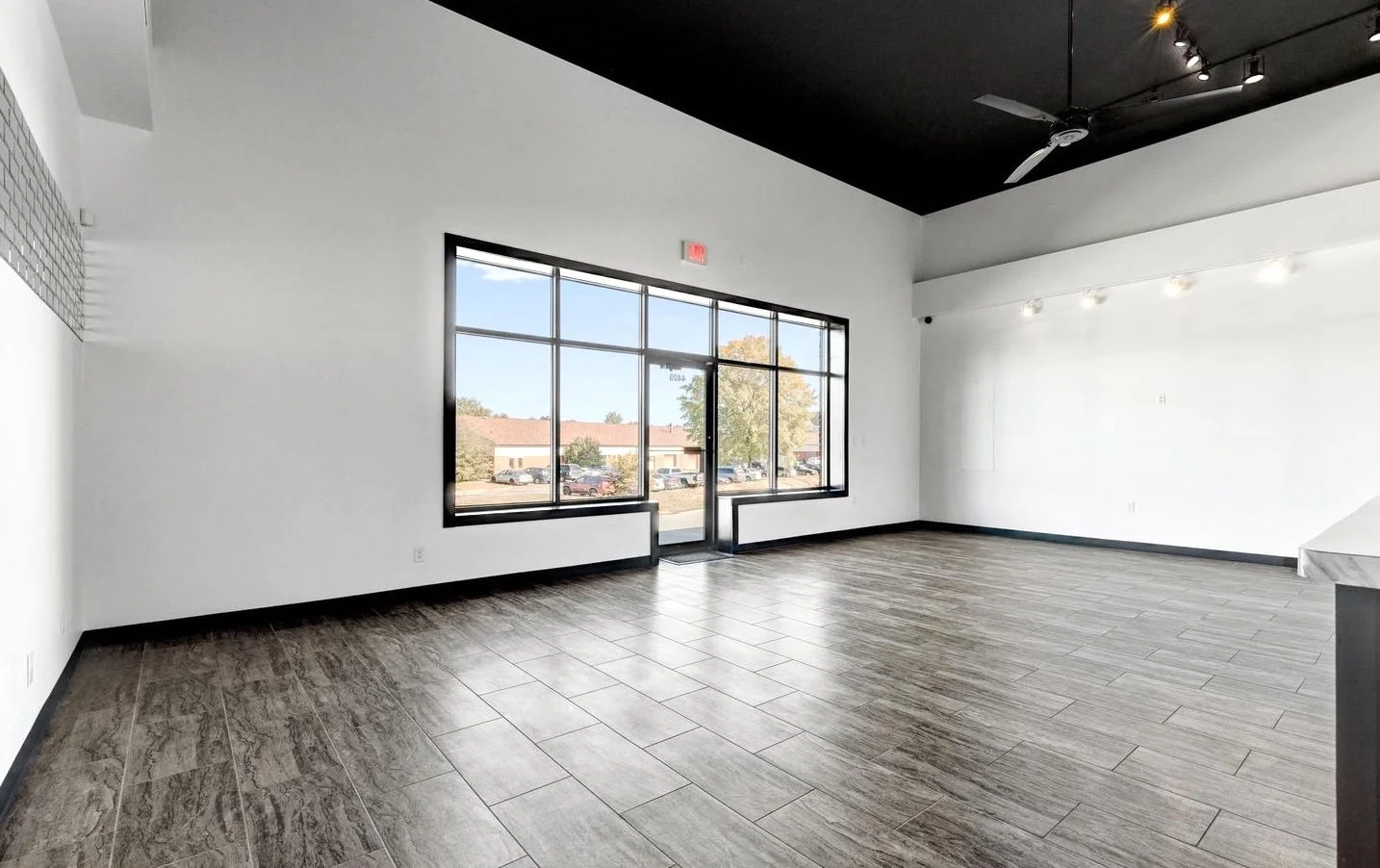 An empty commercial space with large front windows, white walls, wood-look tile flooring, a black ceiling, a ceiling fan, and track lighting.
