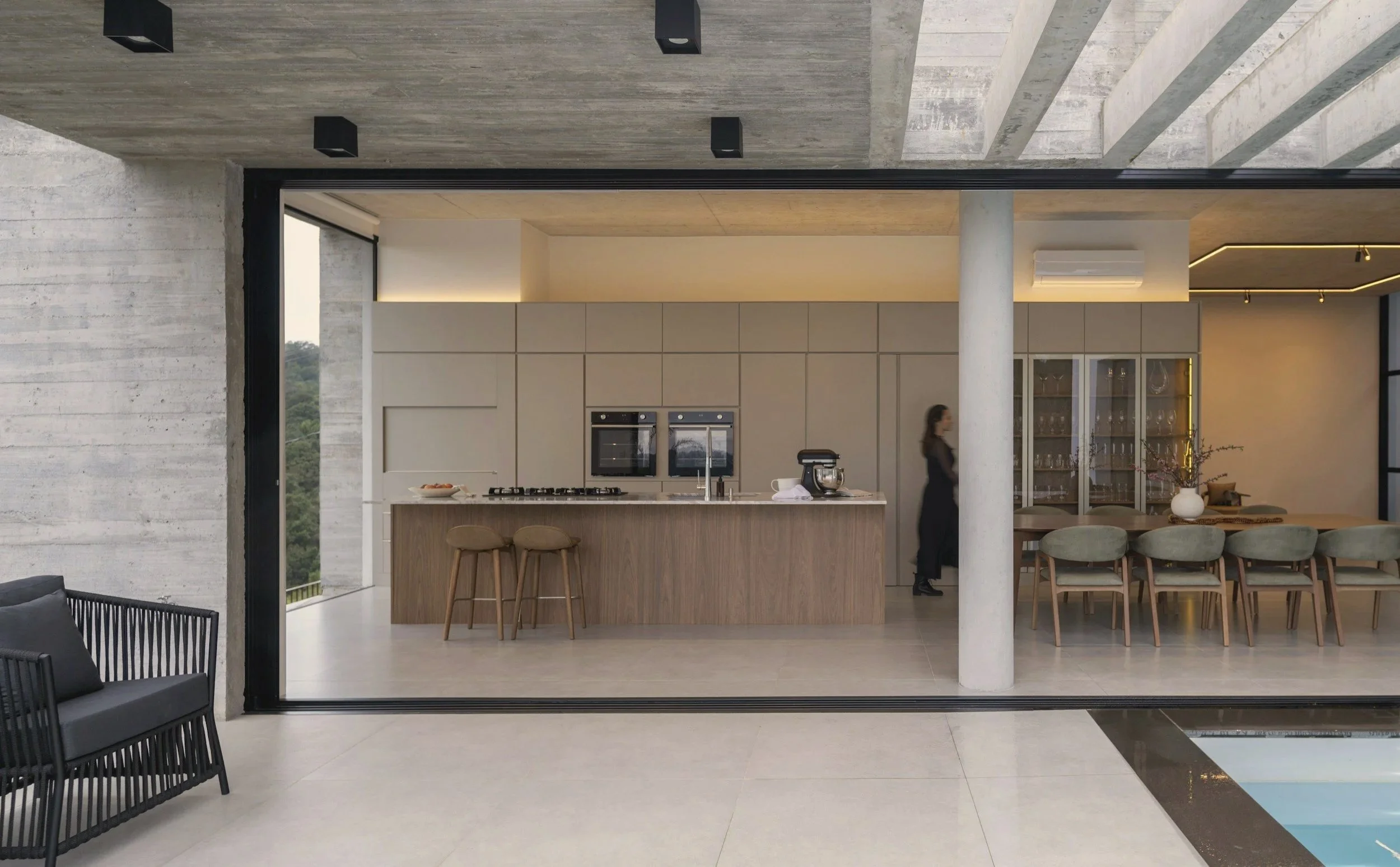 Modern open-concept kitchen and dining area with minimalist design, featuring beige cabinets, a kitchen island with stools, and a dining table with chairs, illuminated by subtle lighting, with a woman standing near the cabinets.