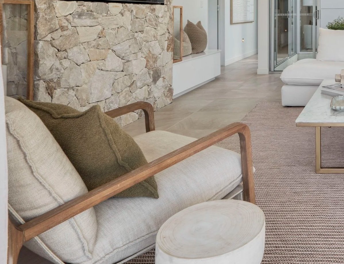 Living room with beige sofa, wooden chair, stone accent wall, glass doors, white and beige decor, and neutral-toned furniture.