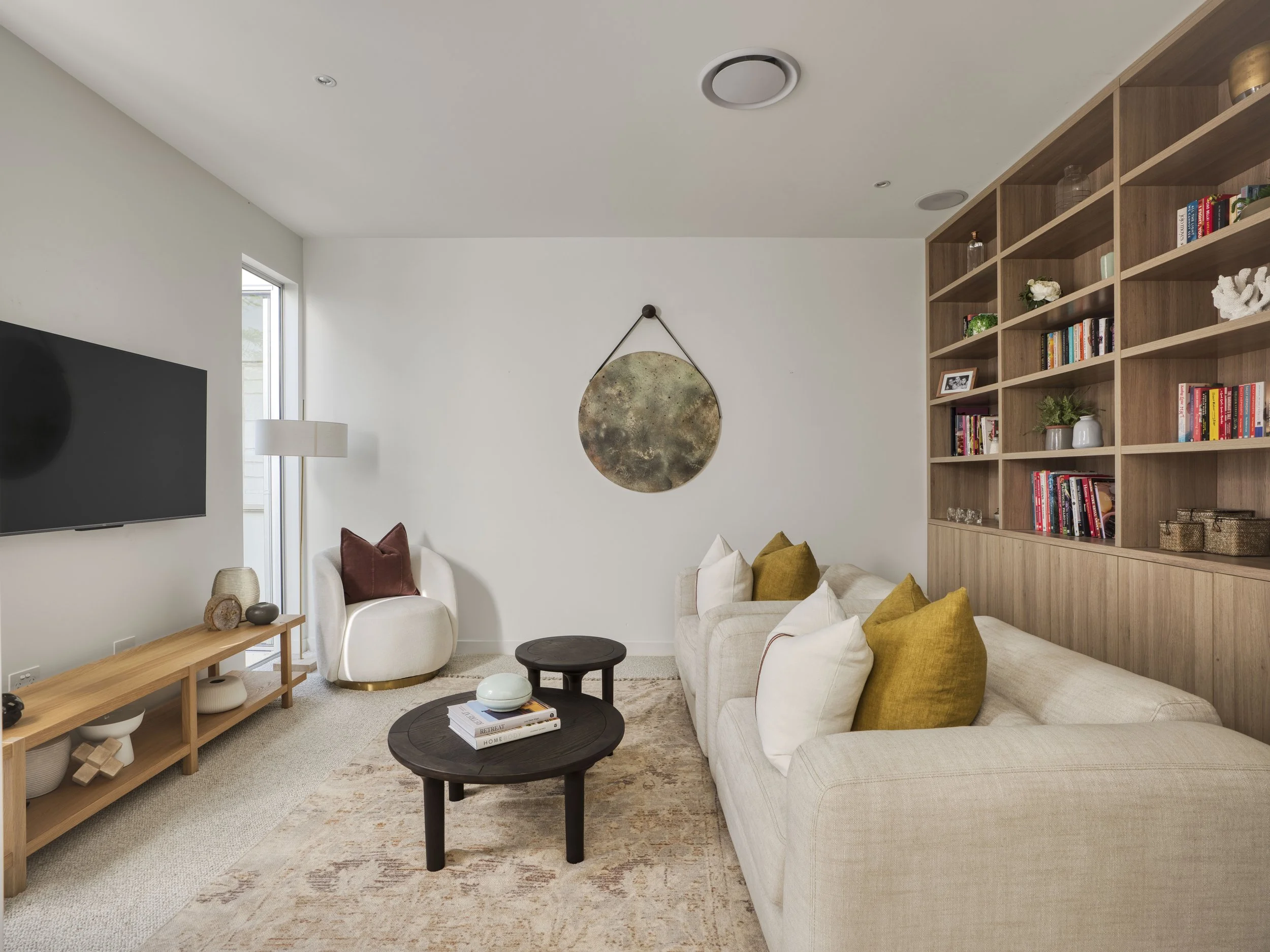 A living room with a beige sofa and yellow pillows, a black coffee table with magazines, a white armchair with a brown pillow, a wooden console with decorative items, a wall-mounted TV, a window, a modern wall hanging, and built-in wooden shelves fil