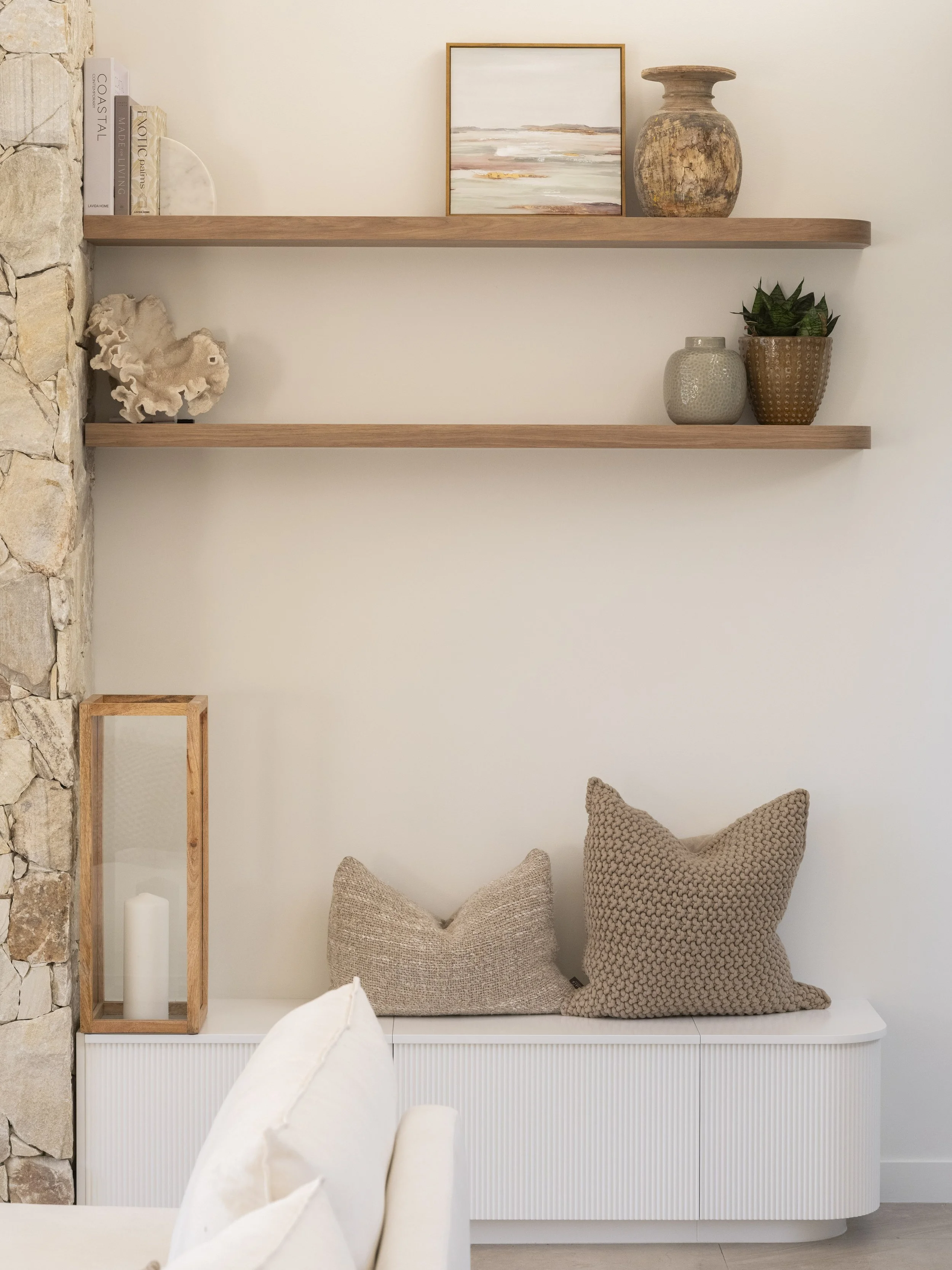 Interior living space with two beige textured pillows on a white cabinet, wooden wall-mounted shelves with decorative items including books, sculptures, a framed landscape painting, and potted plants. A partially visible white couch and a stone wall 