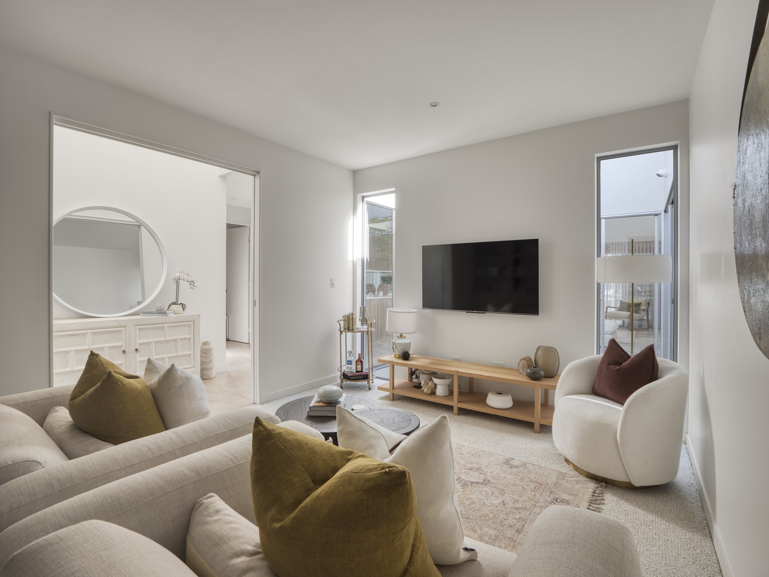 Living room with white walls, large windows, beige sofa with brown pillows, white and wooden furniture, TV on wall, decorative items on coffee table and side table, and a textured wall on the right.