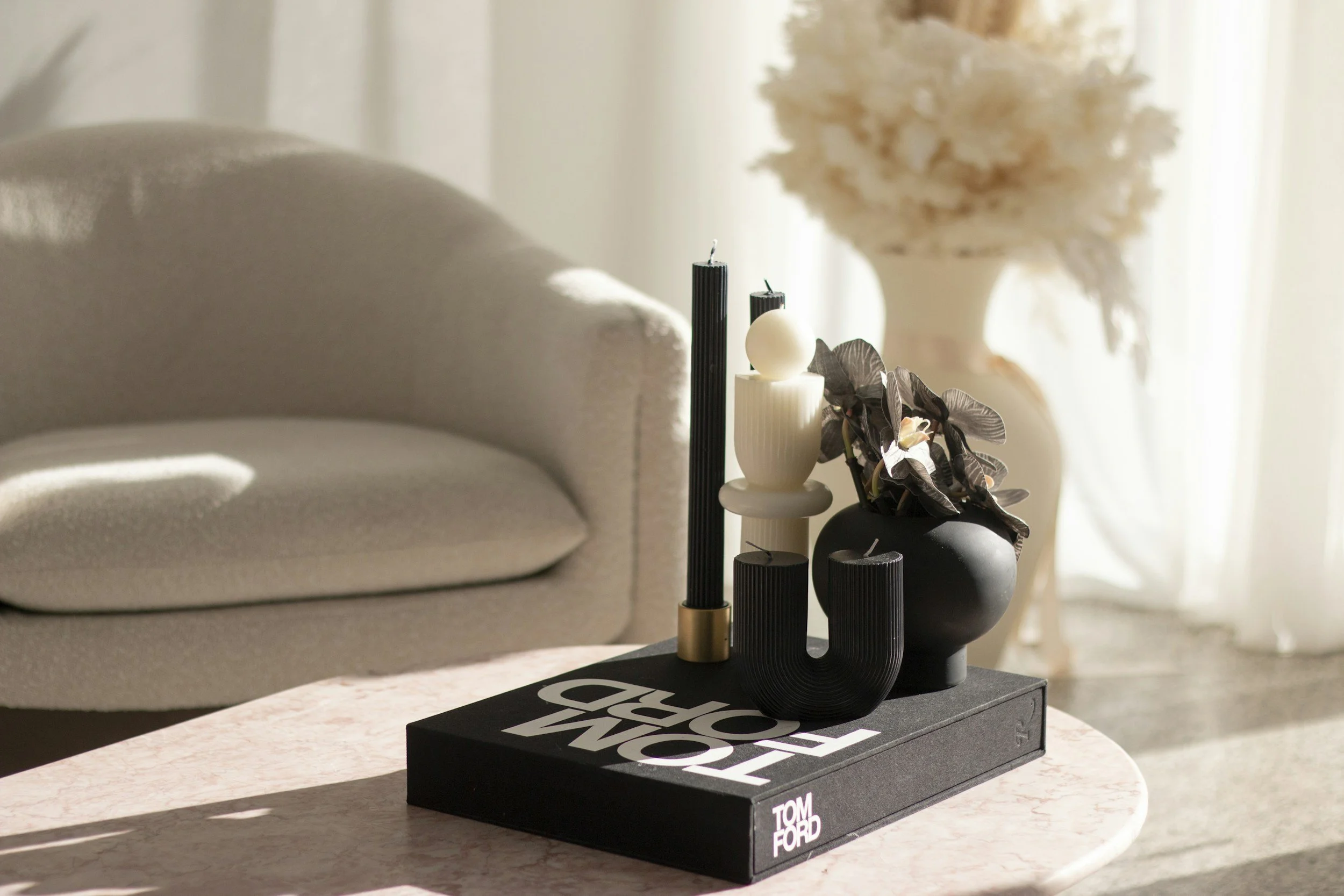 Decorative black and gold candle holder with three candles and foliage, placed on a black box on a light pink marble table, with a beige sofa and a white flower arrangement in the background.