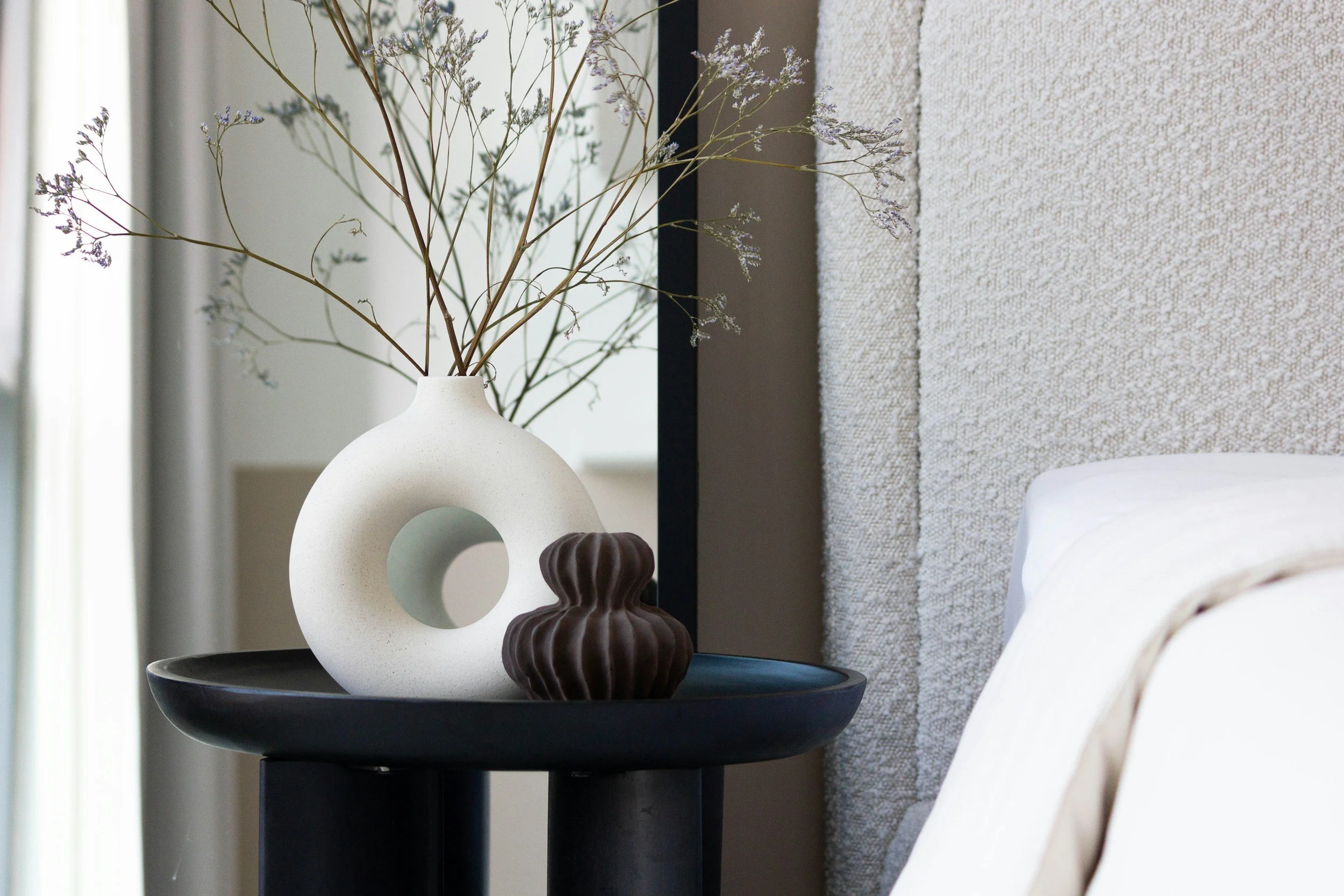 A close-up of a black side table with decorative objects, including a white ceramic vase with dried branches, a small dark brown ribbed ceramic object, and a beige ceramic object with circular cutouts, against a backdrop of a textured beige wall and a white bed.