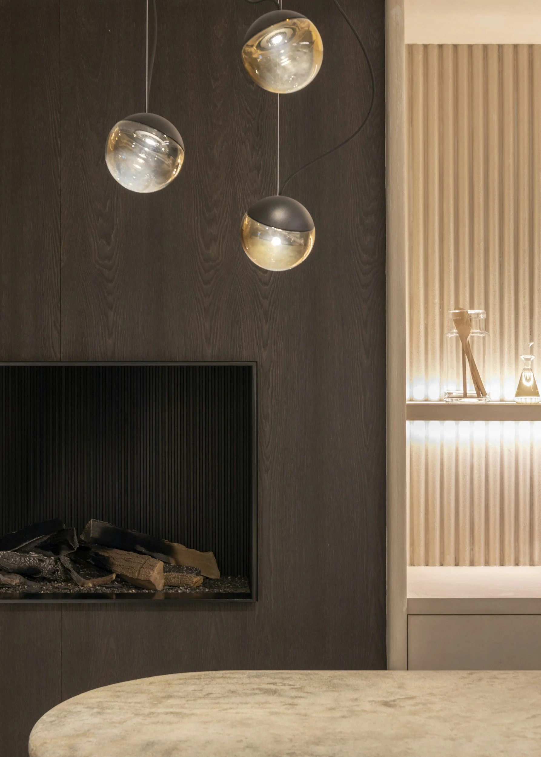 Modern living room with a fireplace containing wood logs, a dark wood panel wall, hanging pendant lights with black shades and glass globes, a beige marble table, and a slatted wooden partition with decorative glass vases.