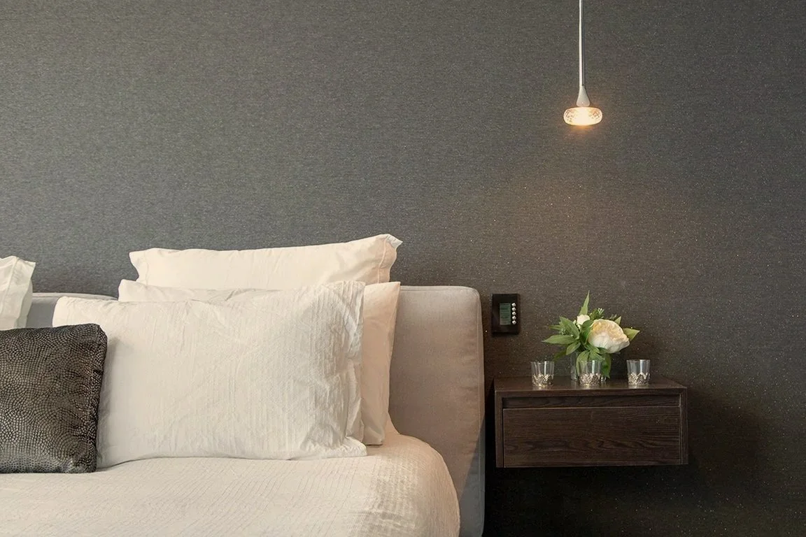 A neatly made bed with white pillows and a dark accent pillow in a modern bedroom, with a floating dark wooden nightstand on a dark textured wall, and a small light pendant hanging above.