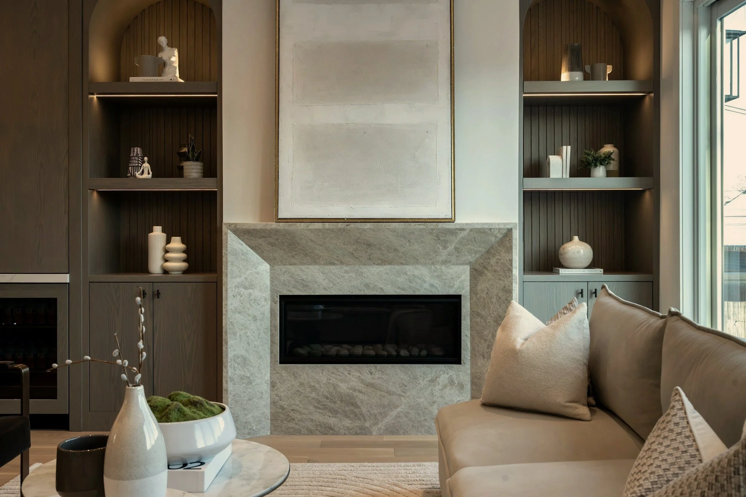 Living room with a beige sofa, beige pillows, a round marble coffee table, a fireplace, and built-in wooden shelves with decorative items.