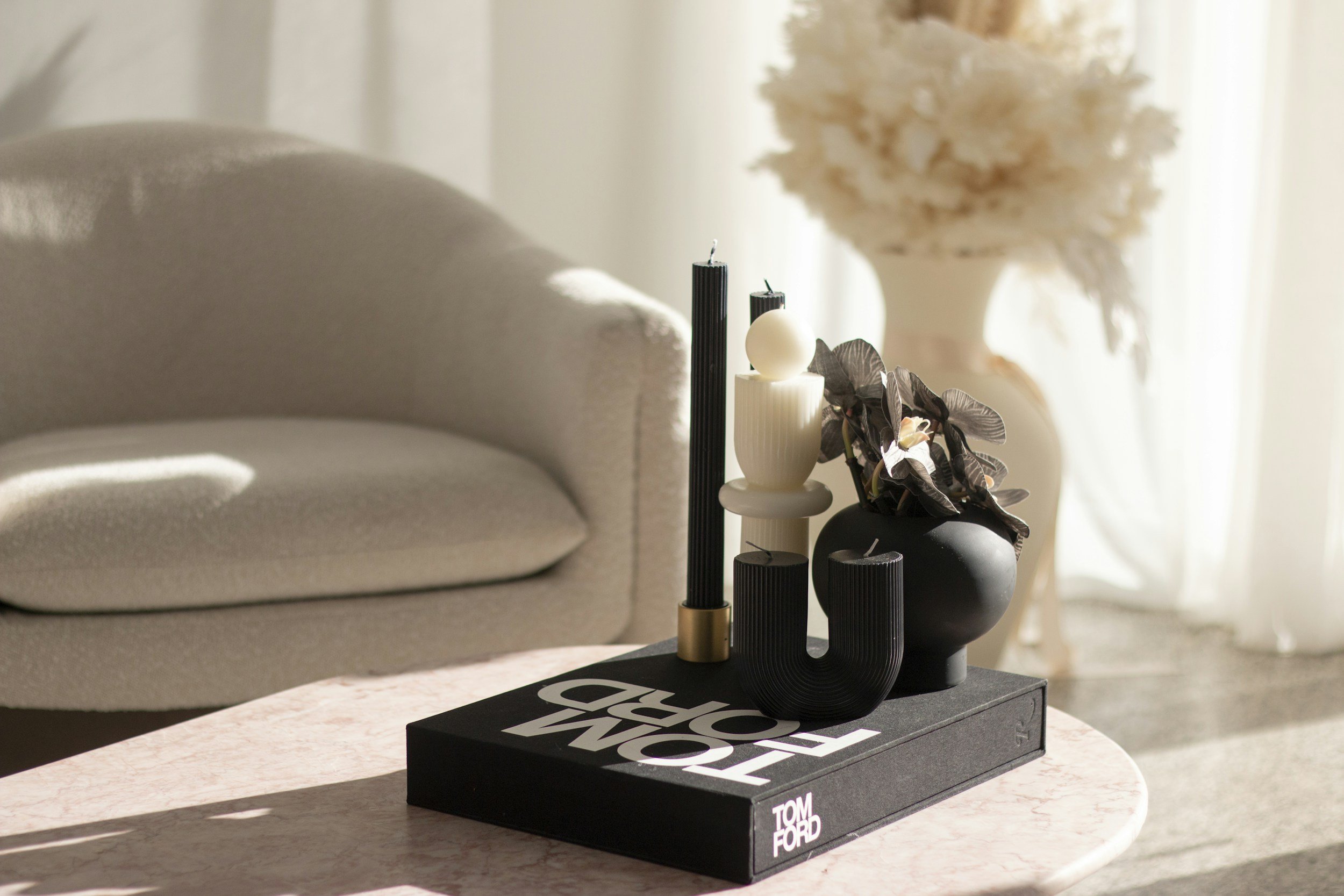 Decorative arrangement with black and white candles, leaves, and a white vase on a black tray, placed on a pink marble table in a softly lit room.
