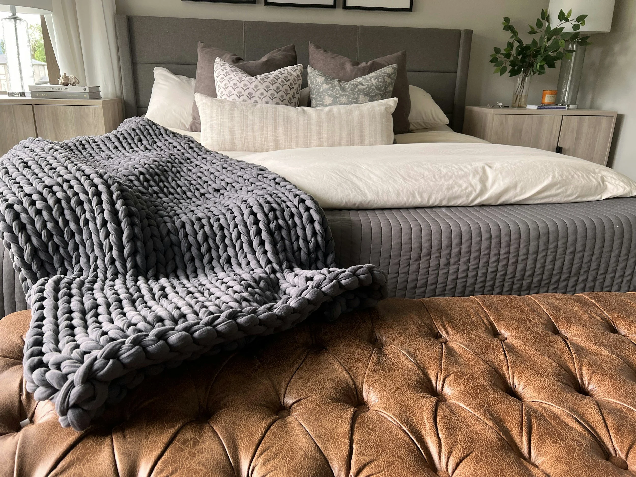 A bed with a variety of decorative pillows, a chunky gray blanket, and a quilted brown leather-style bed frame. There are two nightstands with books, candles, and a glass vase with green foliage, set in a bedroom with framed art on the wall.