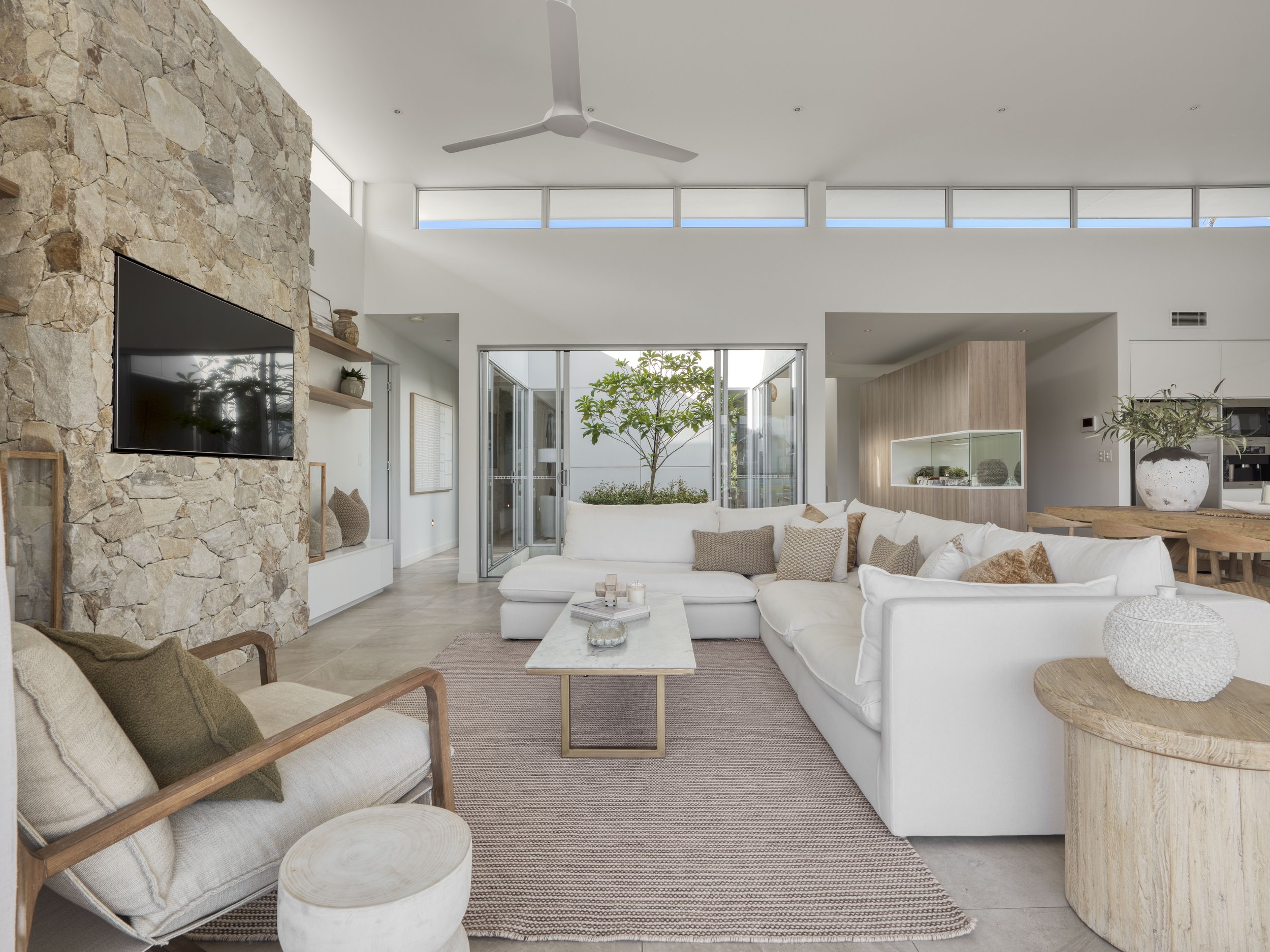 Interior of a modern living room featuring a white sectional sofa with beige pillows, a wooden armchair with cushions, a stone accent wall with mounted flat-screen TV, open glass doors leading to an outdoor space with plants, and high horizontal windows near the ceiling.