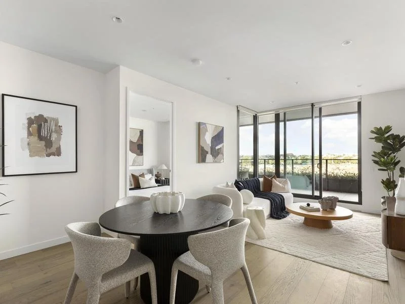 Modern living room with white walls, abstract art, a round dining table with four chairs, a white sofa with cushions, a coffee table, large windows with a balcony view, and potted plants.