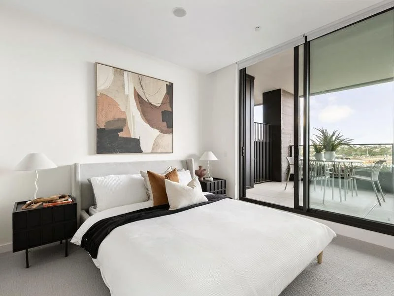 Modern bedroom with a white bed, decorative pillows, abstract artwork above, two bedside lamps, and large glass sliding doors leading to a balcony with outdoor furniture and plants.
