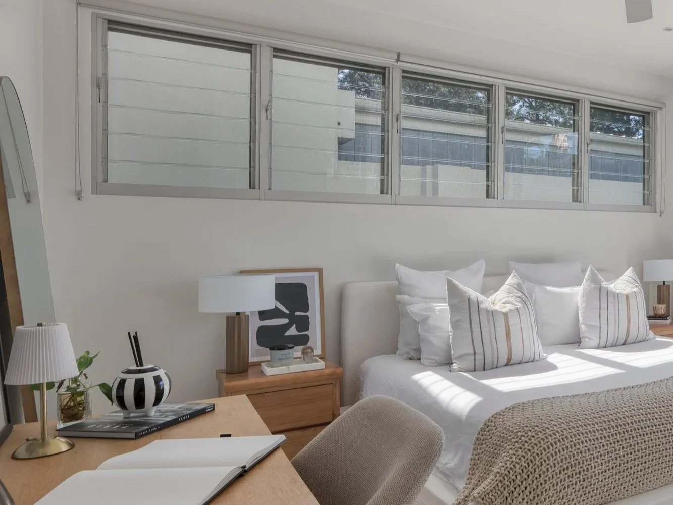 Bedroom with a window, bed with white pillows and a textured beige blanket, wooden nightstand with a lamp and framed art, desk with a book, mirror, and decorative vase