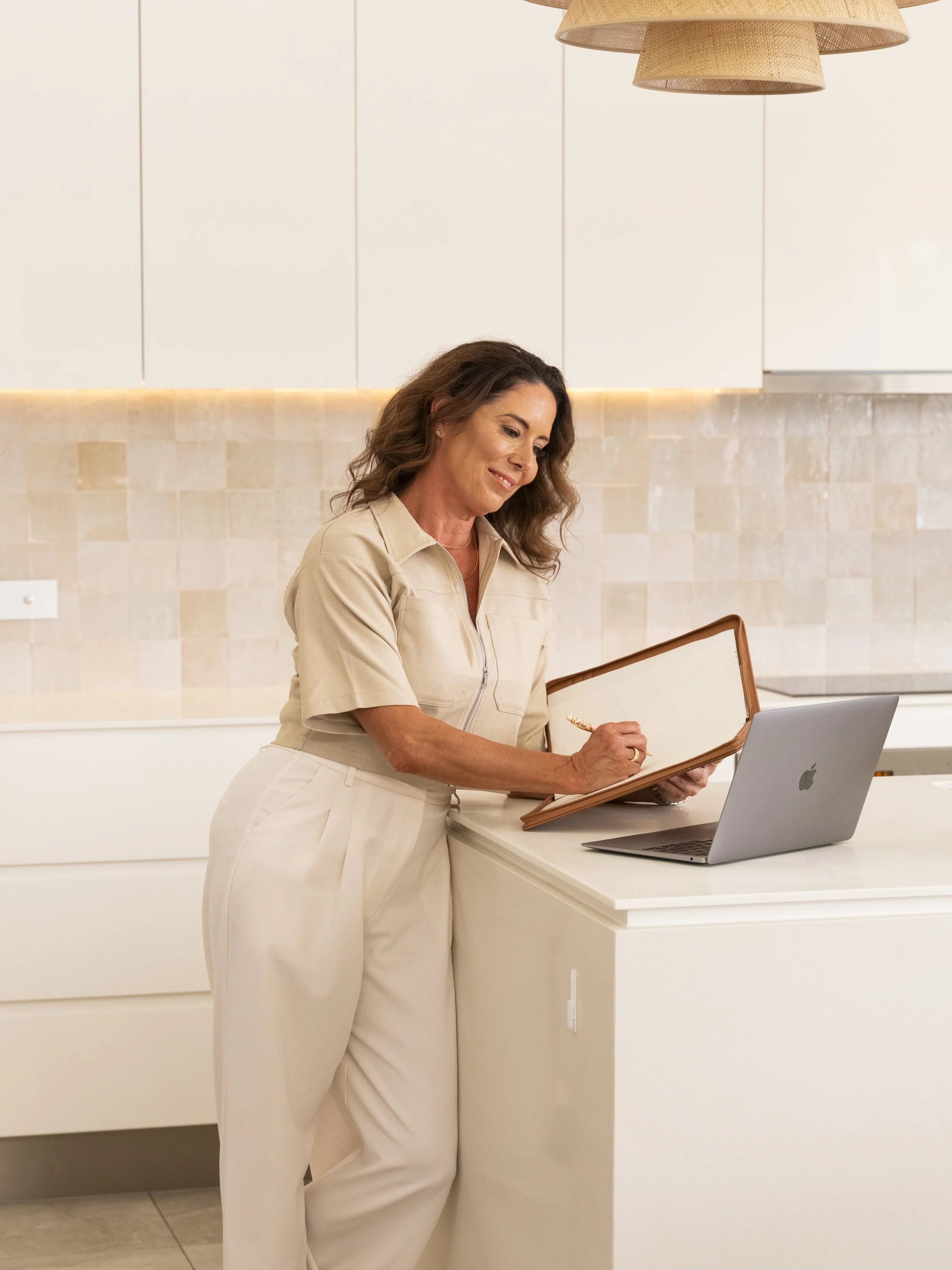 A woman with wavy brown hair looks down at a tablet while standing at a white kitchen counter with a laptop in front of her, in a modern kitchen with beige tiled backsplash and white cabinets.