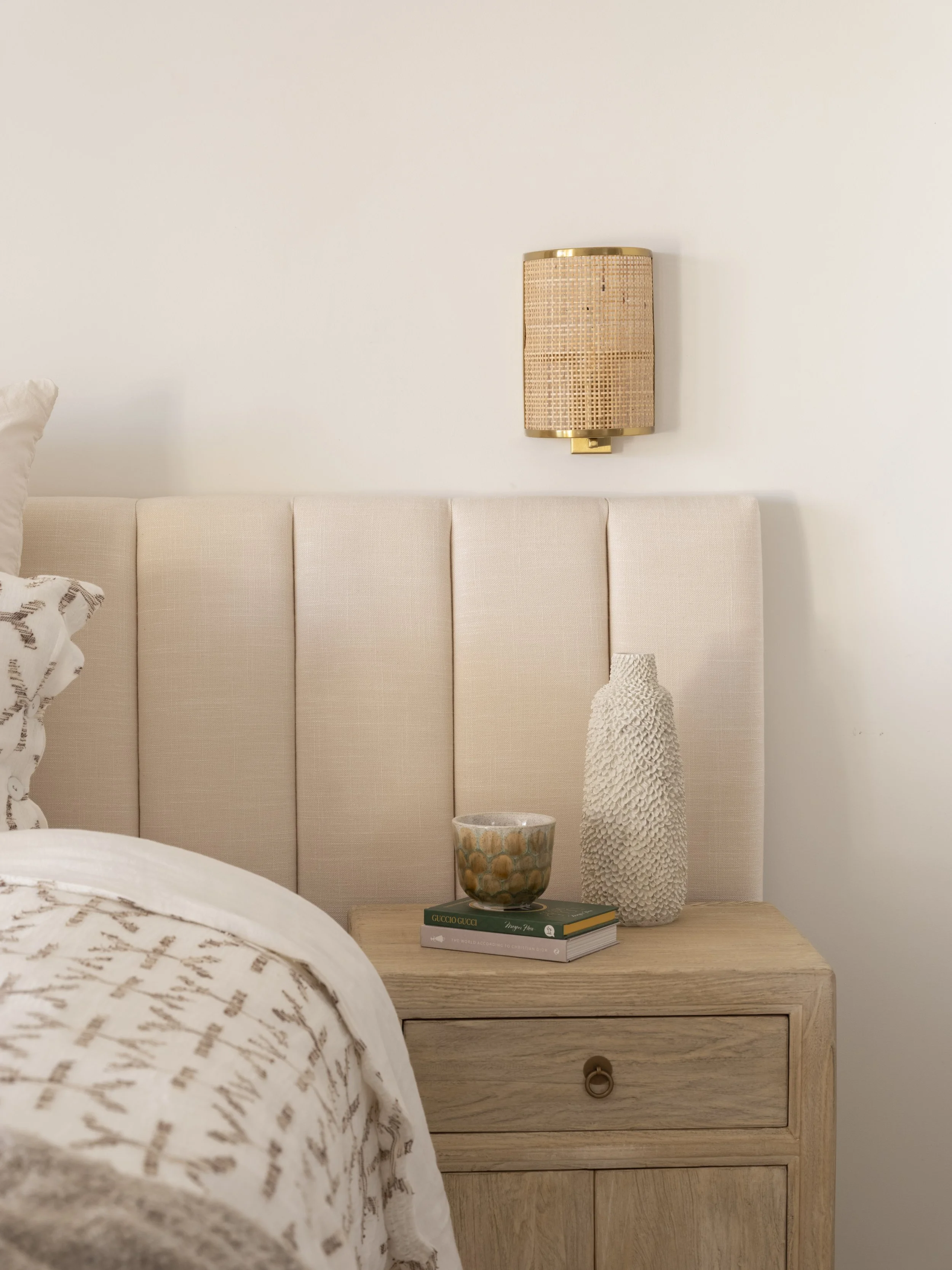 A bedroom scene featuring a beige upholstered headboard, a bedside table with a potted textured white vase, a small bowl, and two books titled 'Gucci Gucci' and another with white and gold accents. Above the headboard is a wall-mounted light fixture 