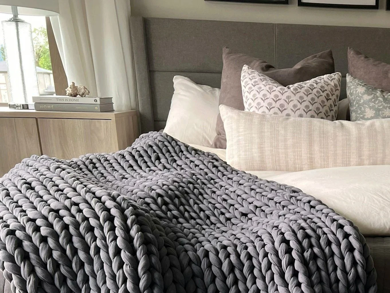 A neatly made bed with a chunky gray knit blanket and various pillows, in front of a gray upholstered headboard in a modern bedroom with a wooden nightstand and decor.