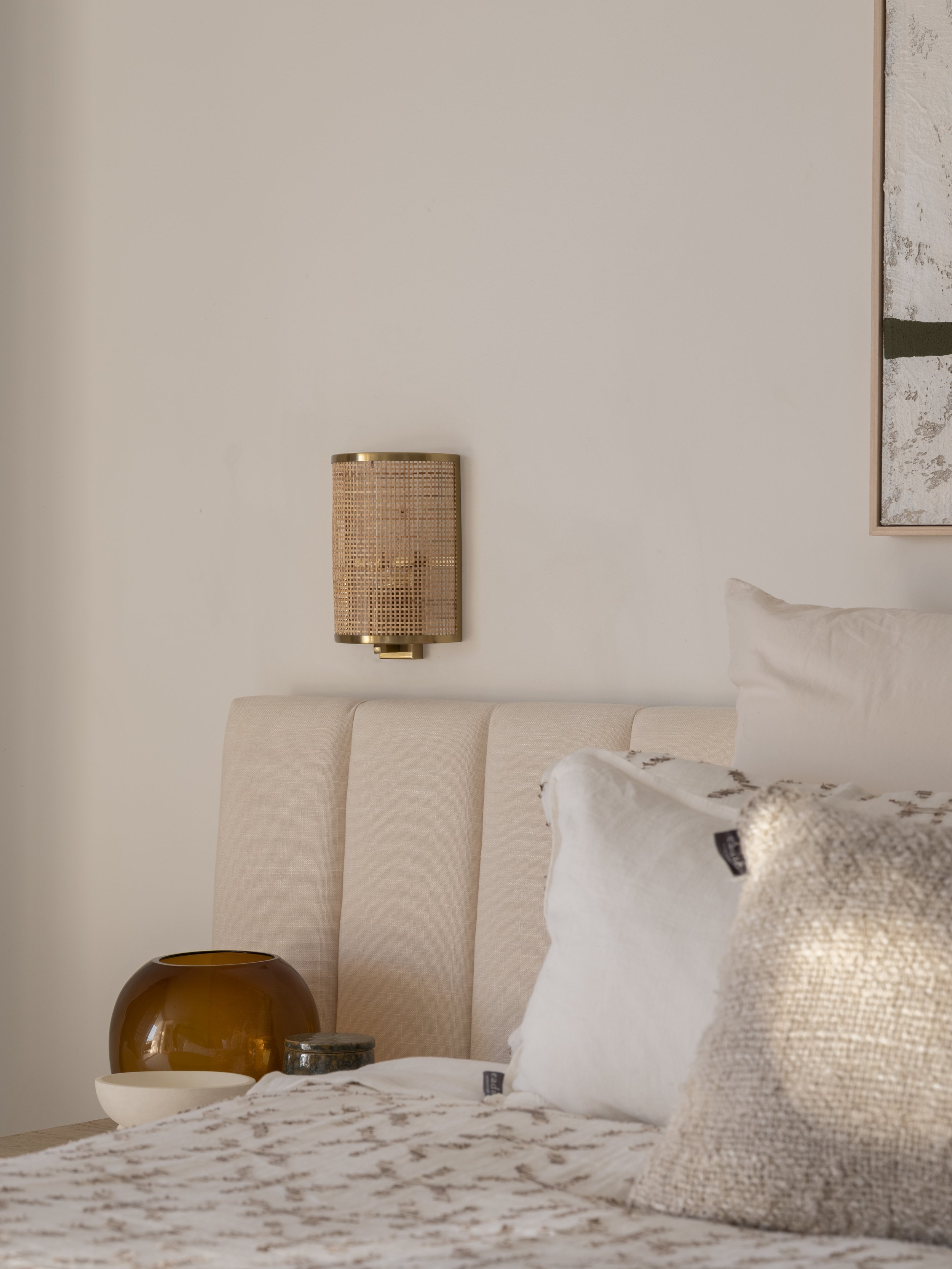 A cozy bedroom corner with a cream-colored upholstered bed, beige and textured pillows, and a decorative textured bedspread. A wooden nightstand holds a large brown vase, a round white bowl, and a small decorative item. A wall-mounted rattan sconce w