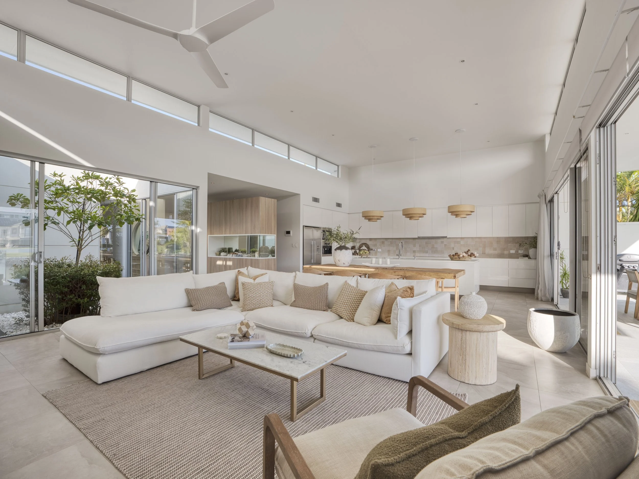 A spacious, modern living room with large glass sliding doors, white walls, and high ceilings. The room is decorated with a large white sectional sofa with beige pillows, a small coffee table with books and decor, and a green indoor plant. There are 