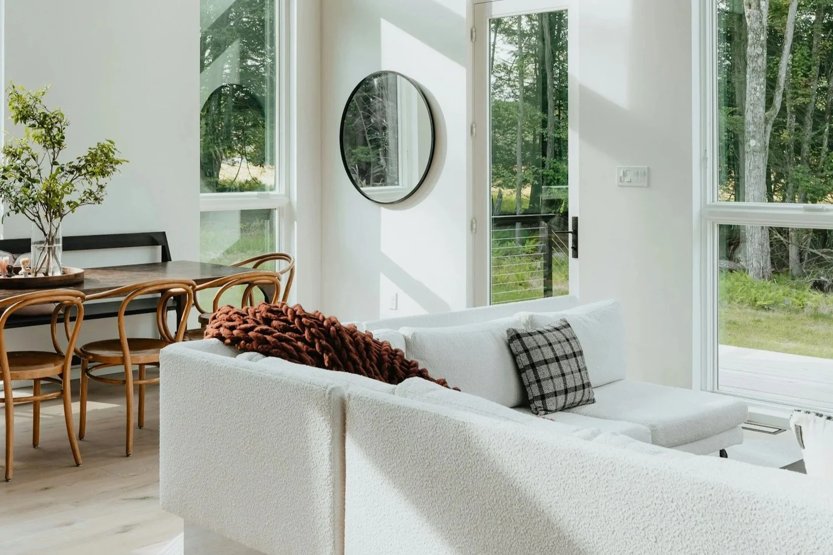 A bright and airy living room with large windows and a glass door leading outside to a wooded area. It features a white sofa with a plaid pillow and a brown chunky throw blanket, a wooden dining table with matching chairs, and a wall mirror.