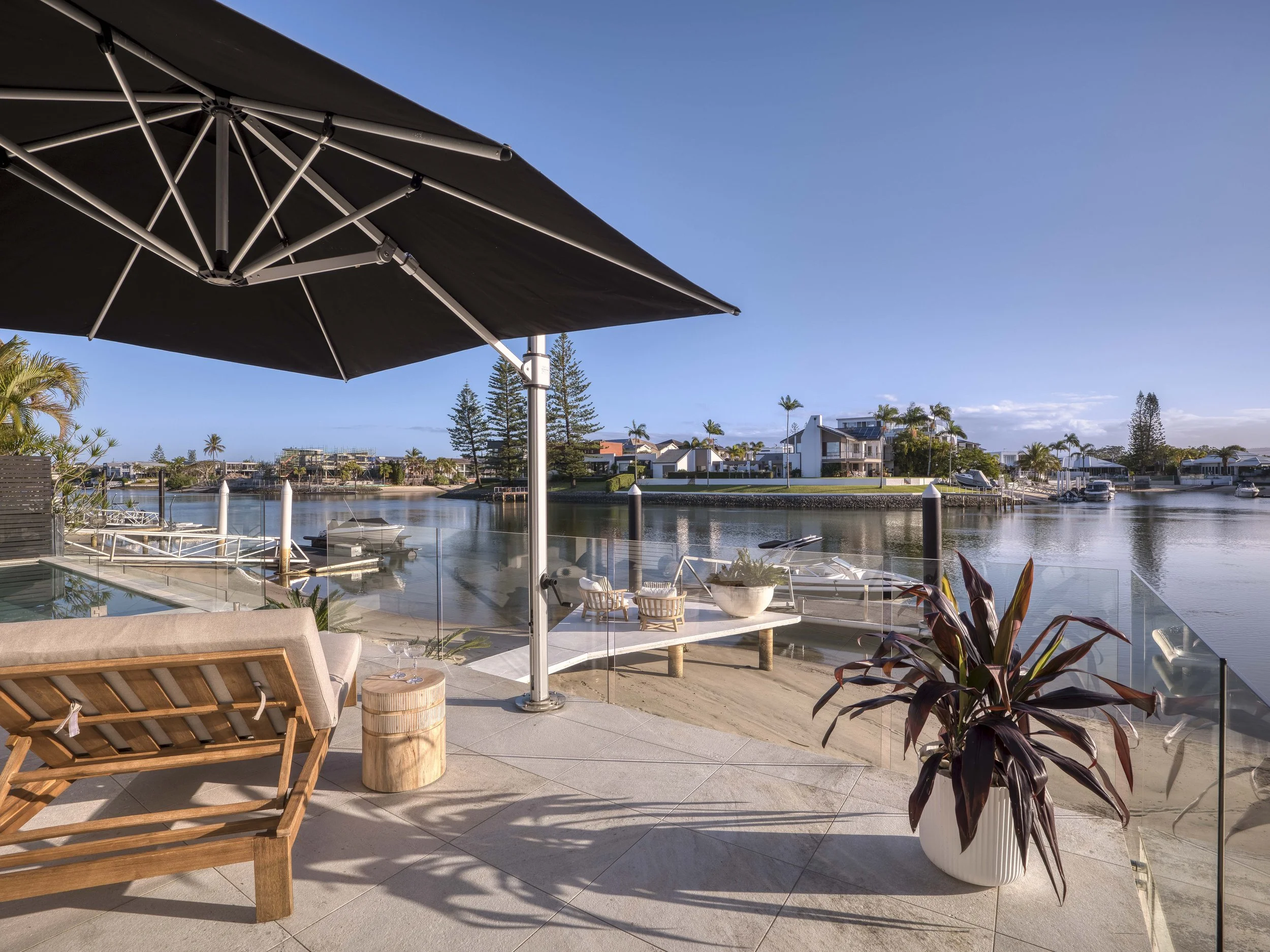 A view from a balcony overlooking a waterway with modern houses, palm trees, and boats, under a blue sky with some clouds. There is a black patio umbrella in the foreground, alongside outdoor furniture and plants.