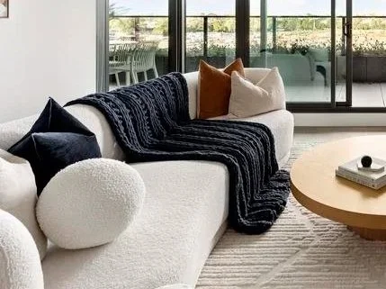 Modern living room with a white sofa, black and tan throw pillows, a black throw blanket, a round wooden coffee table, and large windows with a view of a patio.