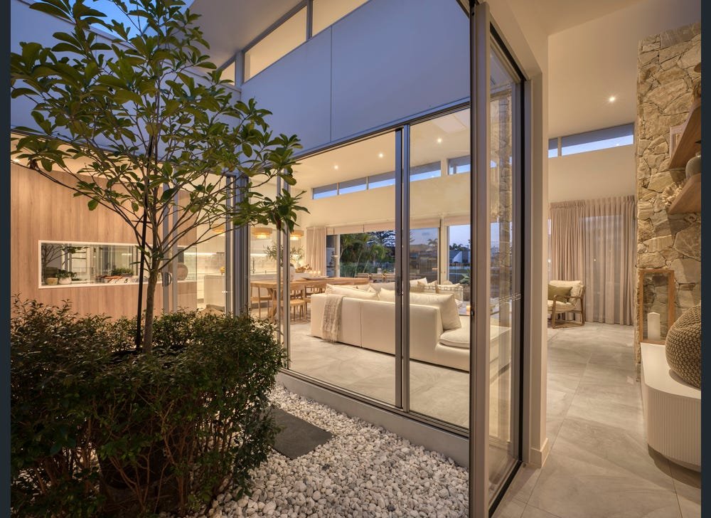 Modern indoor living room with large glass windows, white sofas, and a dining area, seen from the outdoor patio with small white rocks and plants.