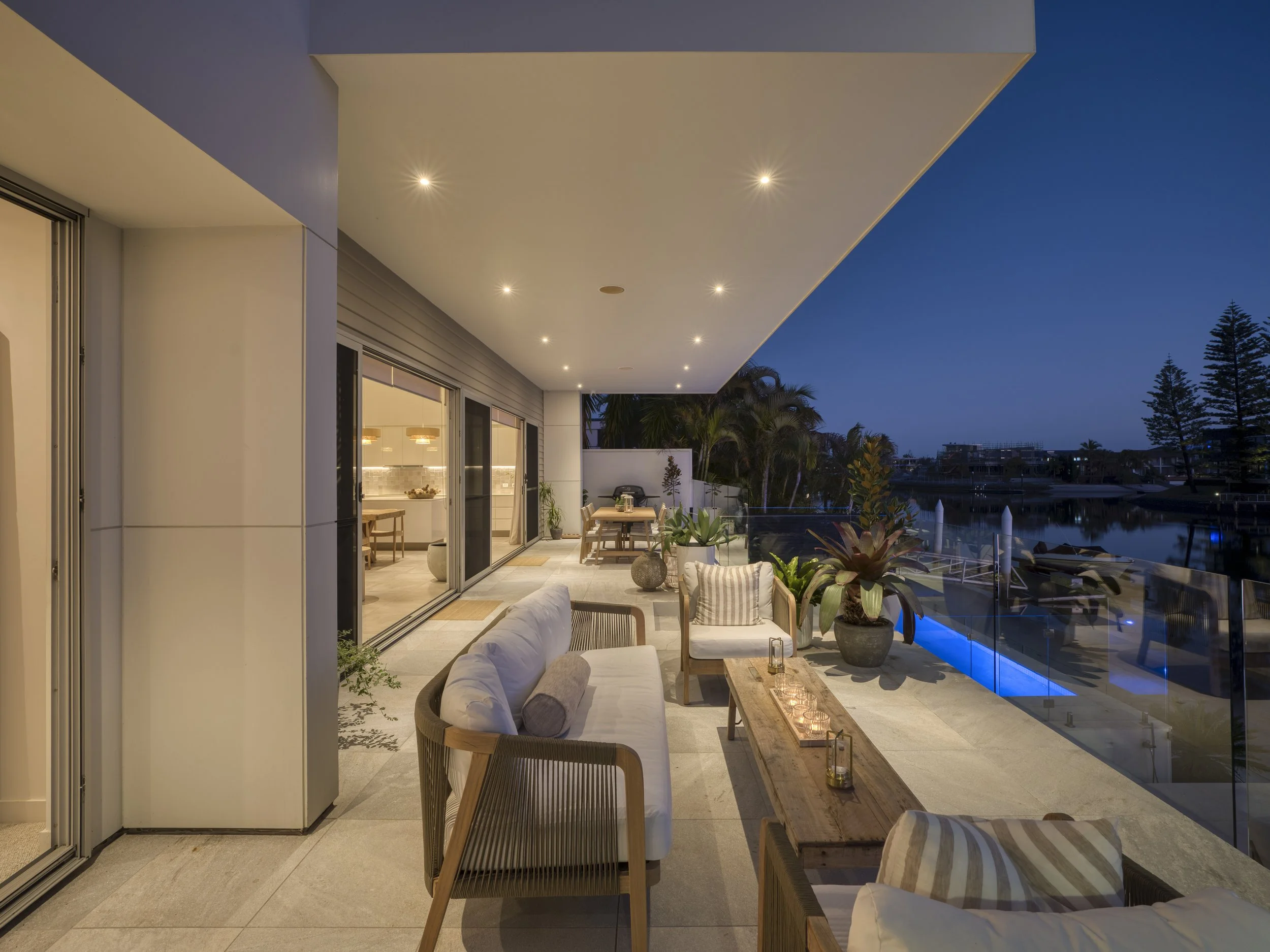 Nighttime view of an outdoor patio with furniture, potted plants, and a swimming pool, illuminated by embedded ceiling lights, overlooking a cityscape with trees and buildings.