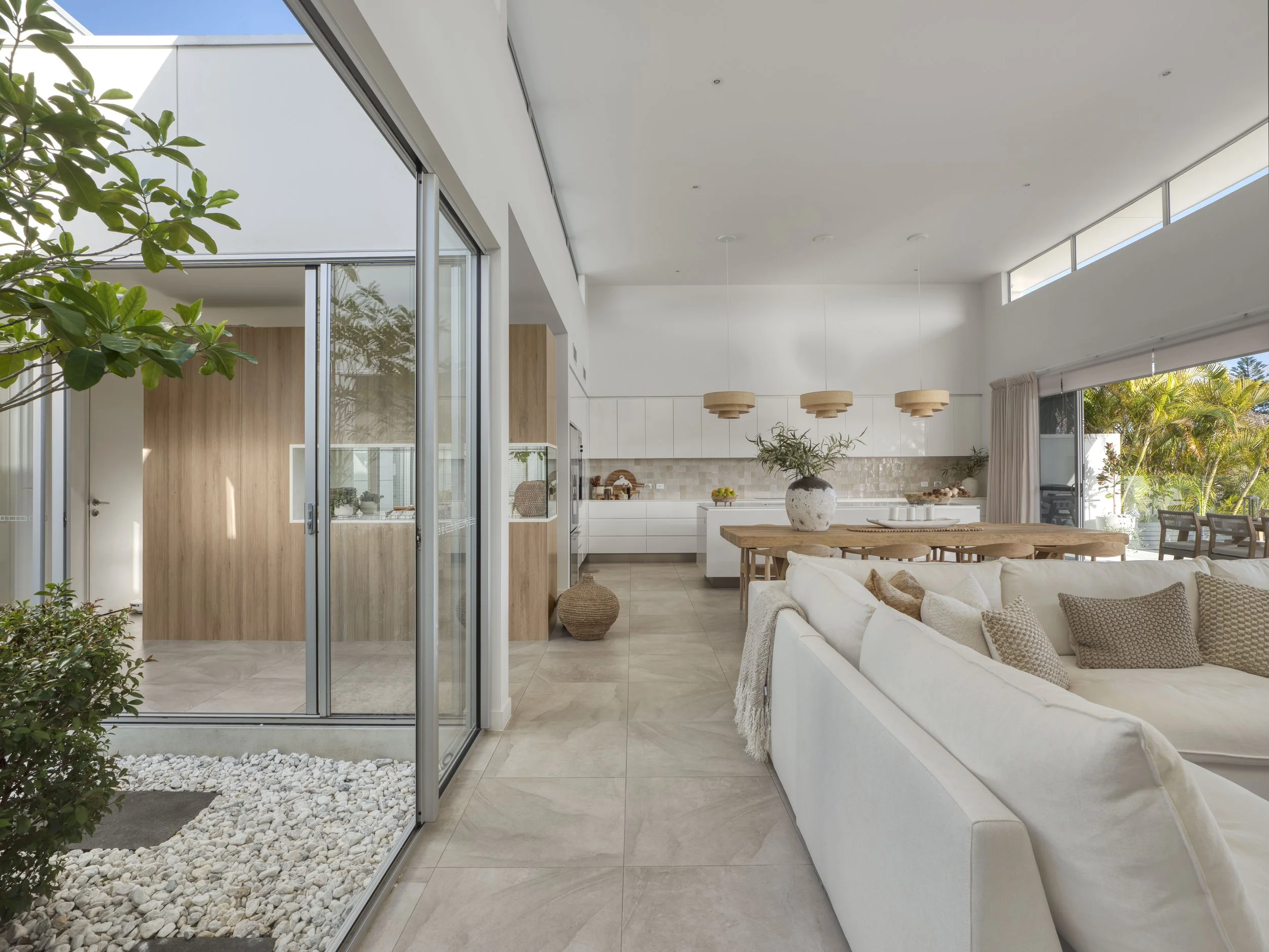 Modern living room with white sofa, wooden dining table, hanging pendant lights, large glass windows, and indoor plants.