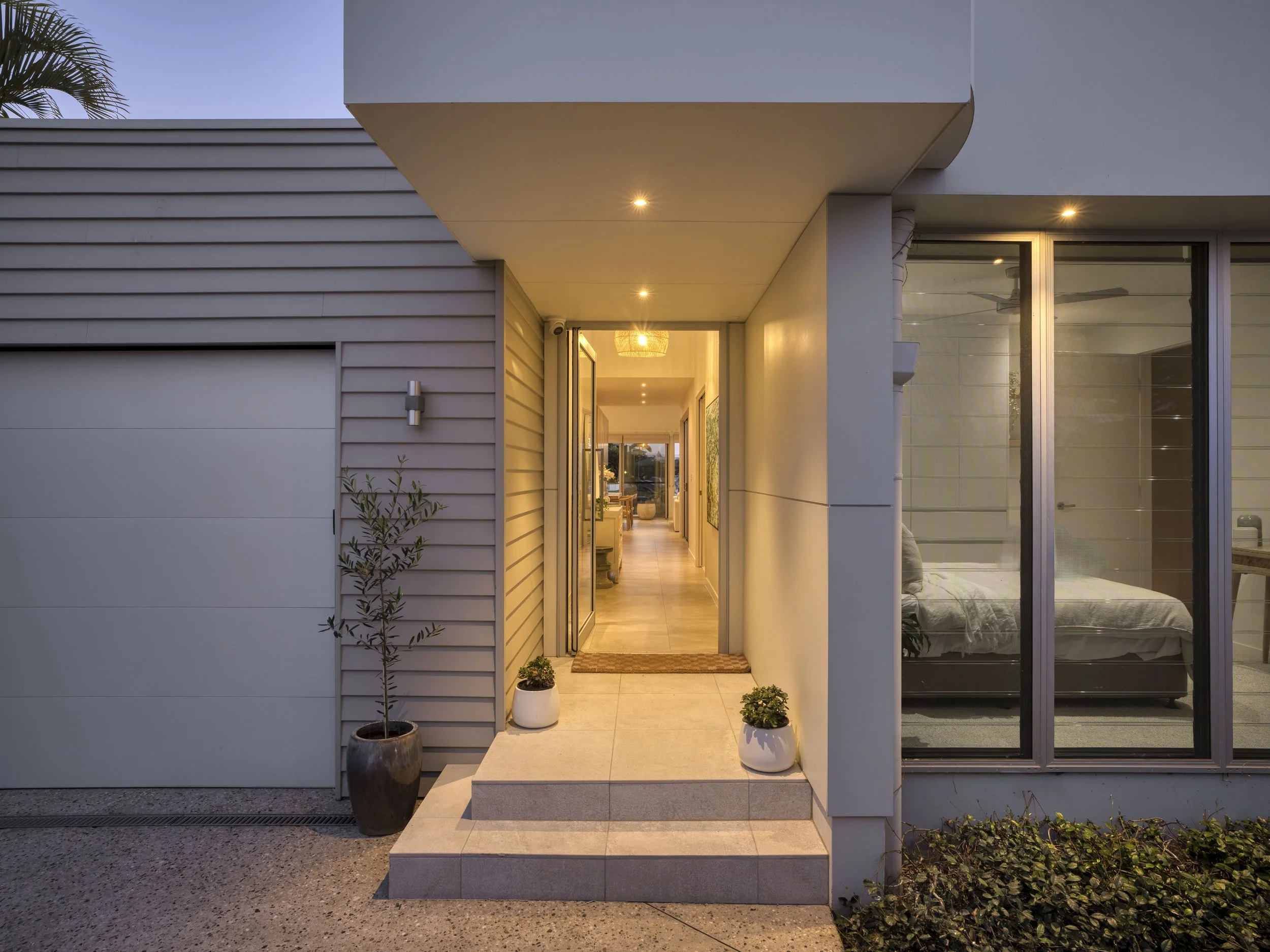 The entrance to a modern house with a porch, potted plants, and interior visible through glass doors and windows.