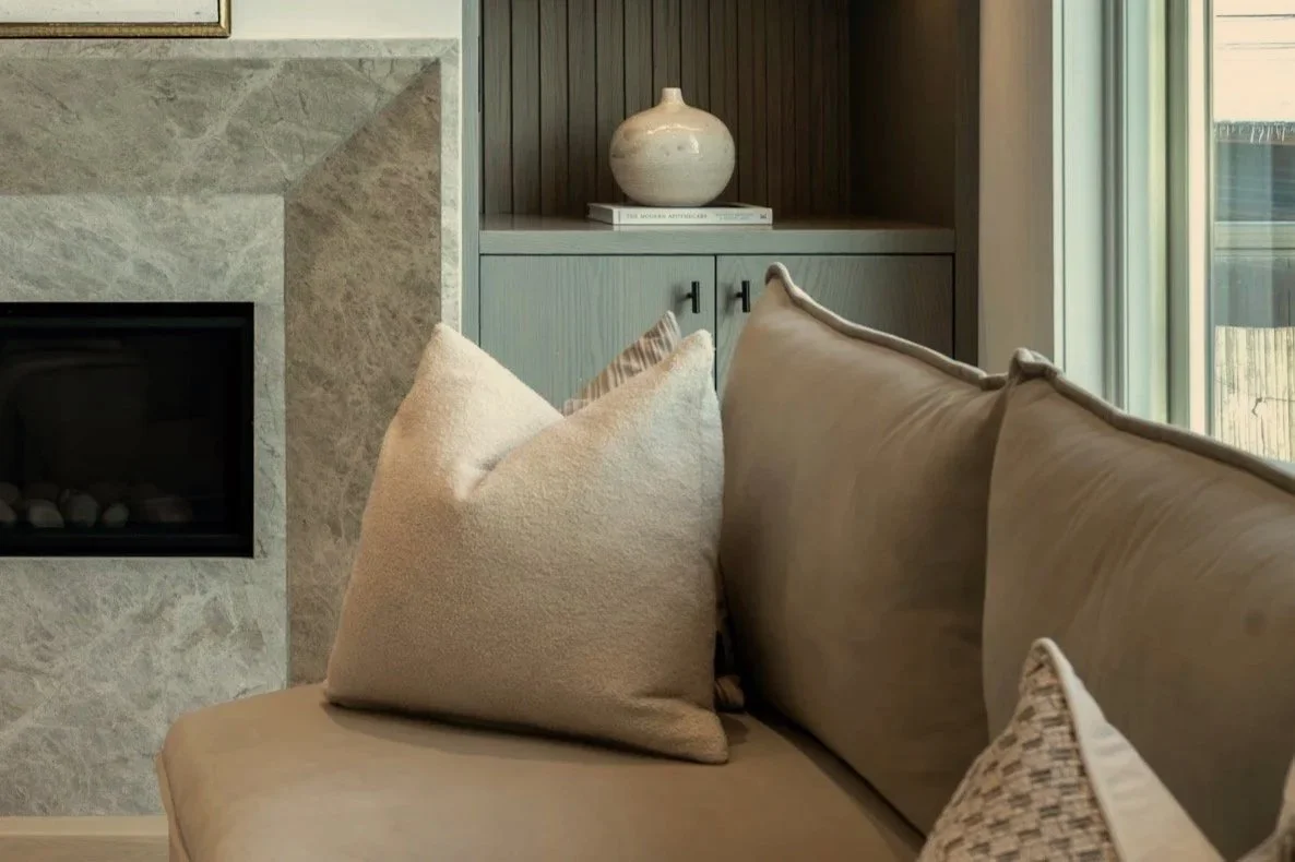 A beige couch with various pillows in front of a fireplace with a stone border, and a built-in cabinet with a decorative vase and books, next to a large window.