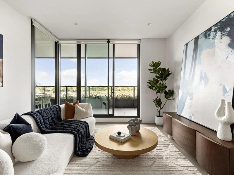 Modern living room with large glass sliding doors leading to a balcony, white walls, a white sofa with black and brown pillows, a black blanket, a round wooden coffee table with decor, a large abstract art piece on the wall, a potted plant, and a pat