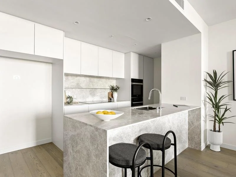 Modern white kitchen with a marble island, black bar stools, and a potted plant in a white planter.