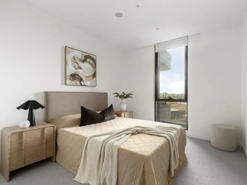 Bedroom with beige bed, black pillows, wooden nightstands, black table lamp, artwork above bed, and large window showing city view