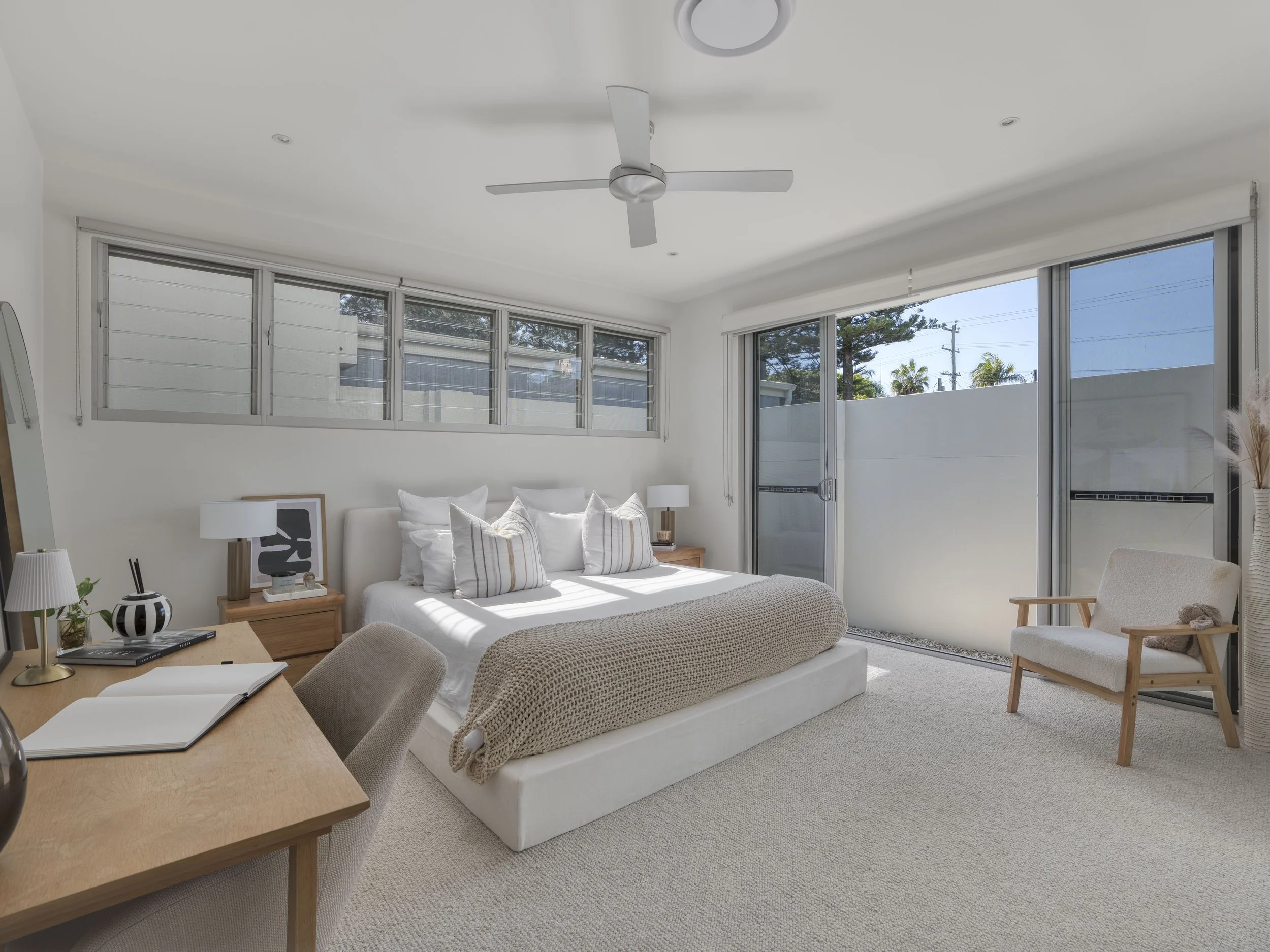 Living room with white walls, large windows, a white bed with pillows, a wooden table with an open notebook, beige chairs, and a ceiling fan.