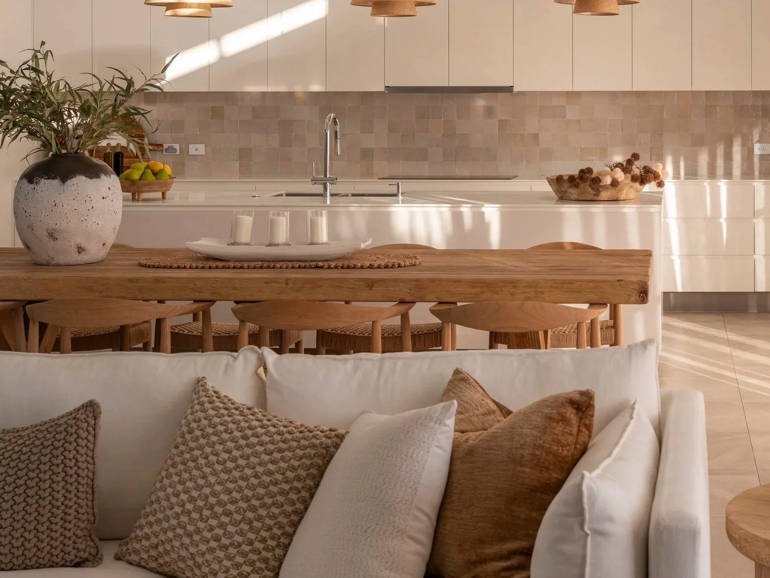 Modern kitchen with a light-colored backsplash, wooden dining table and chairs, decorative vase with greenery, candle holder, and a sofa with beige cushions in the foreground.