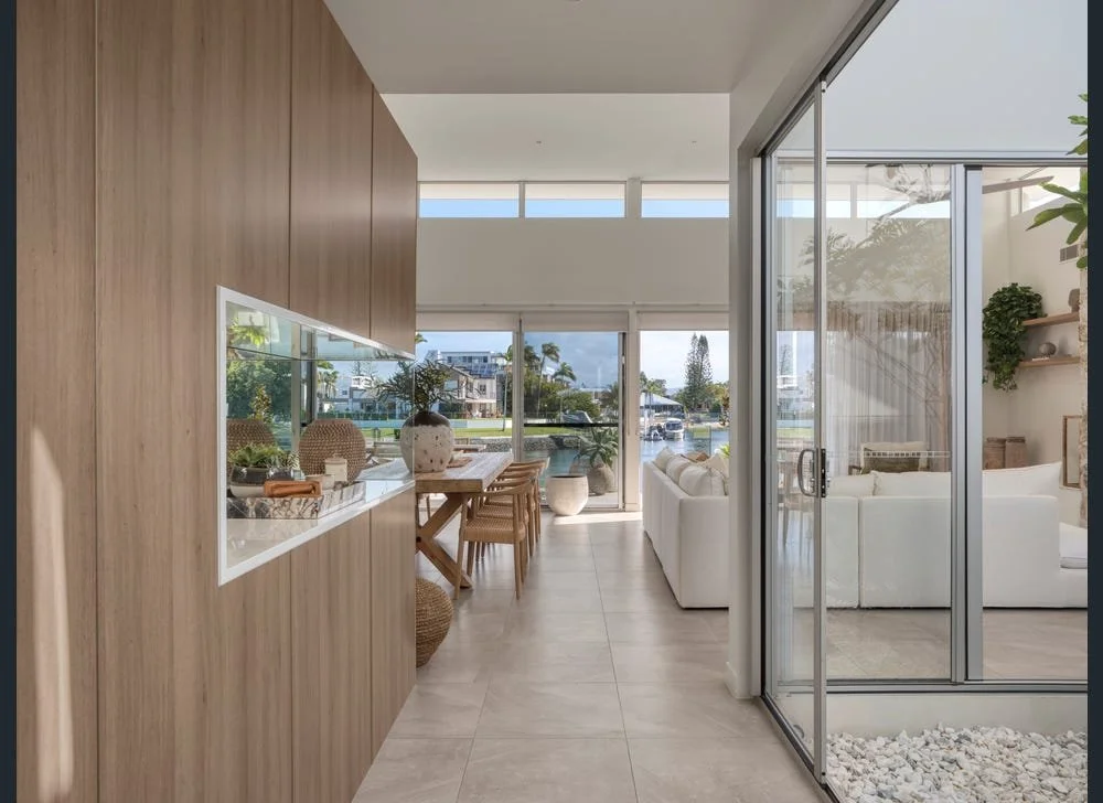 Interior view of a modern living space with a wooden accent wall, a built-in window display, and a large sliding glass door leading to an outdoor patio with white furniture, plants, and a view of a canal and buildings outside.