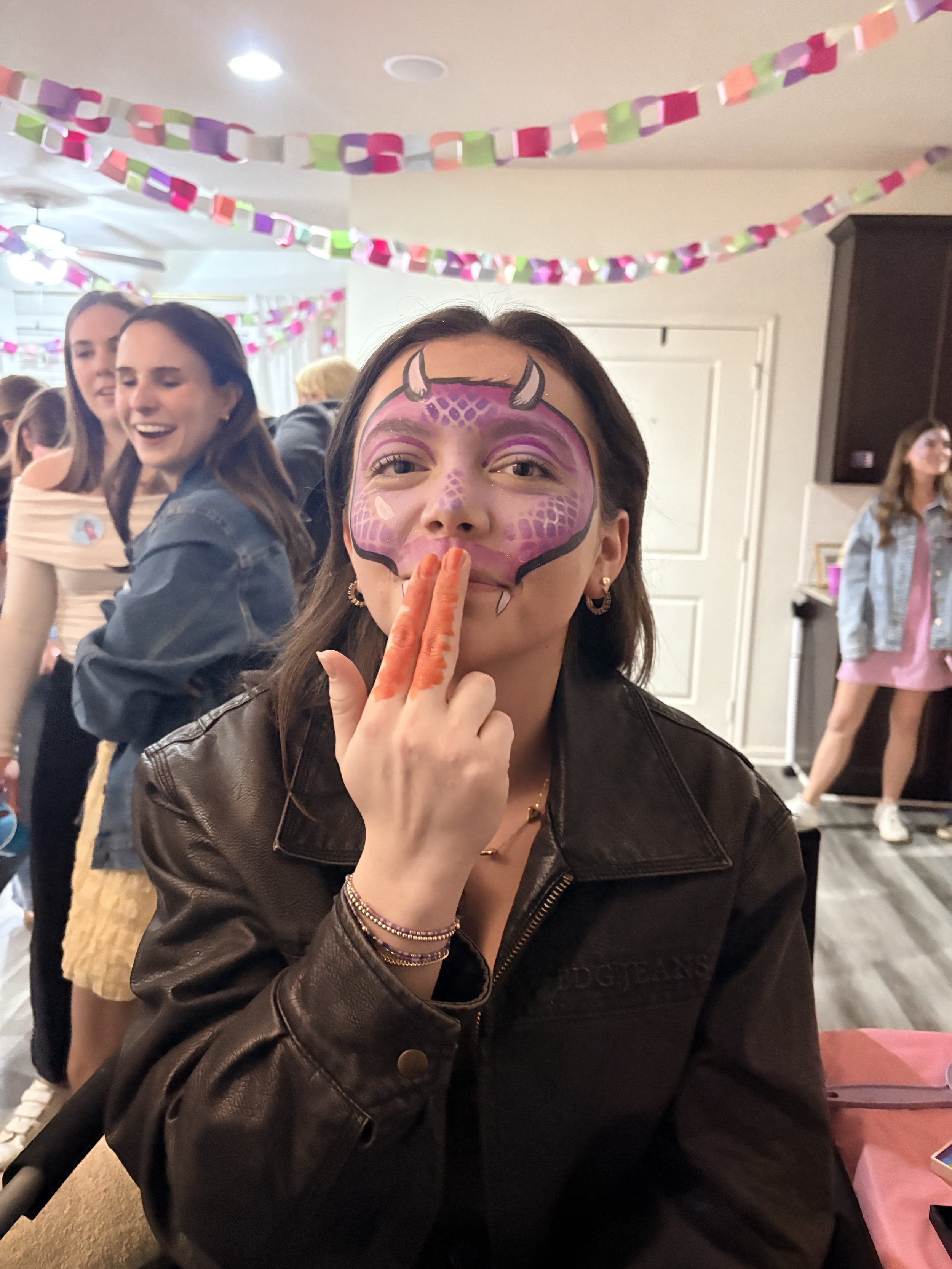 Girl with face painted as a purple dragon with horns, making a shh gesture at a birthday party, decorated with colorful paper chains.