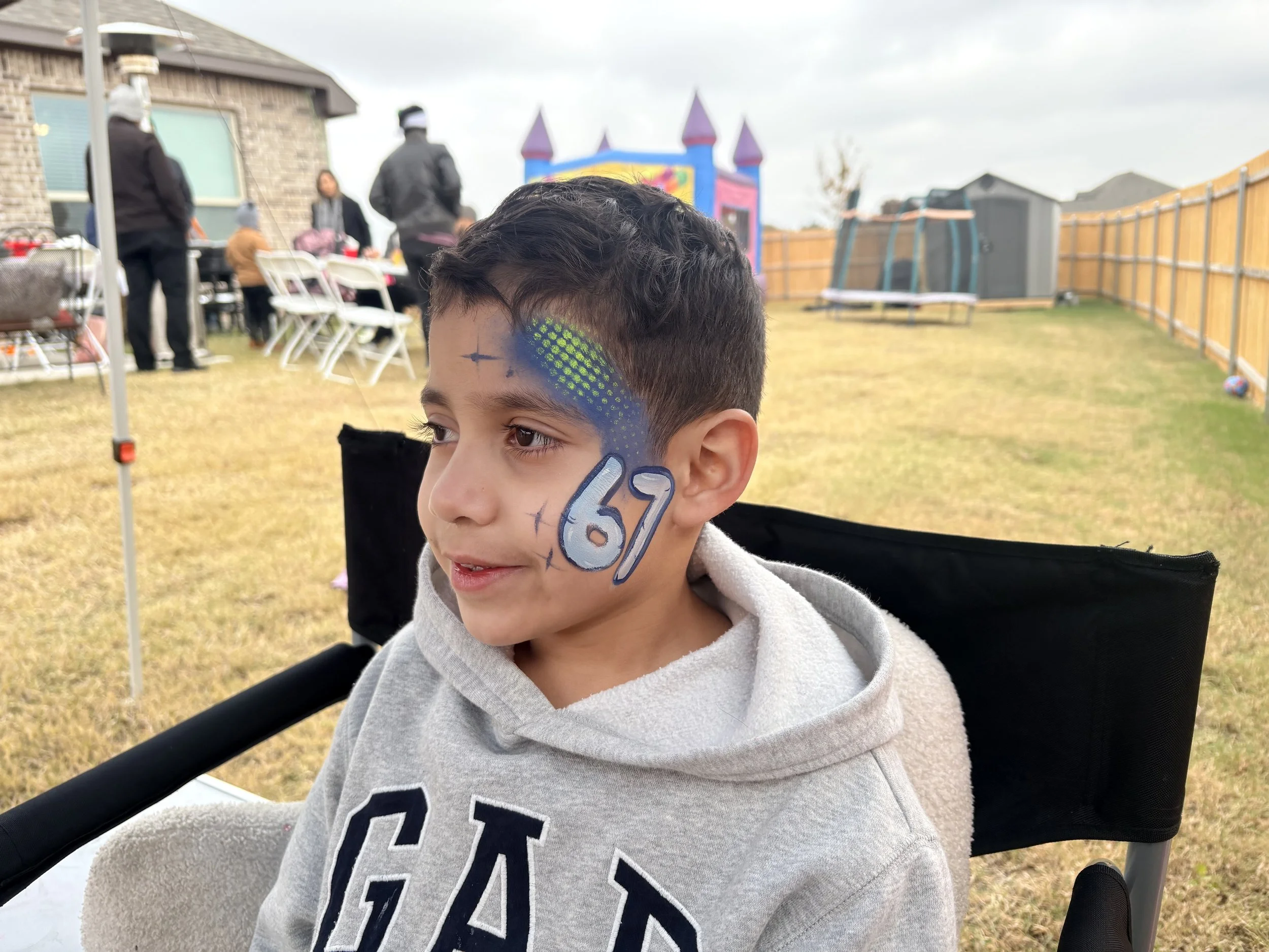 Young boy with face paint that reads '67' at outdoor birthday party with tables, chairs, and a bounce house in the background.