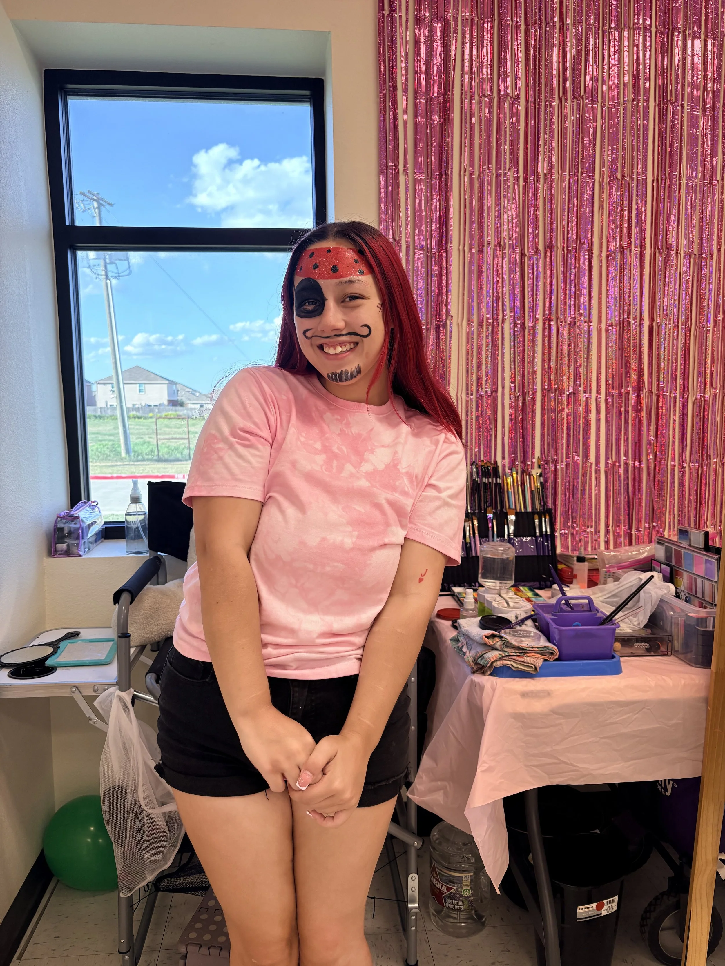 A young woman with face paint in the style of a ladybug, smiling while standing in a room with makeup supplies and a pink sequin curtain. She has red hair, black shorts, and a pink tie-dye shirt.