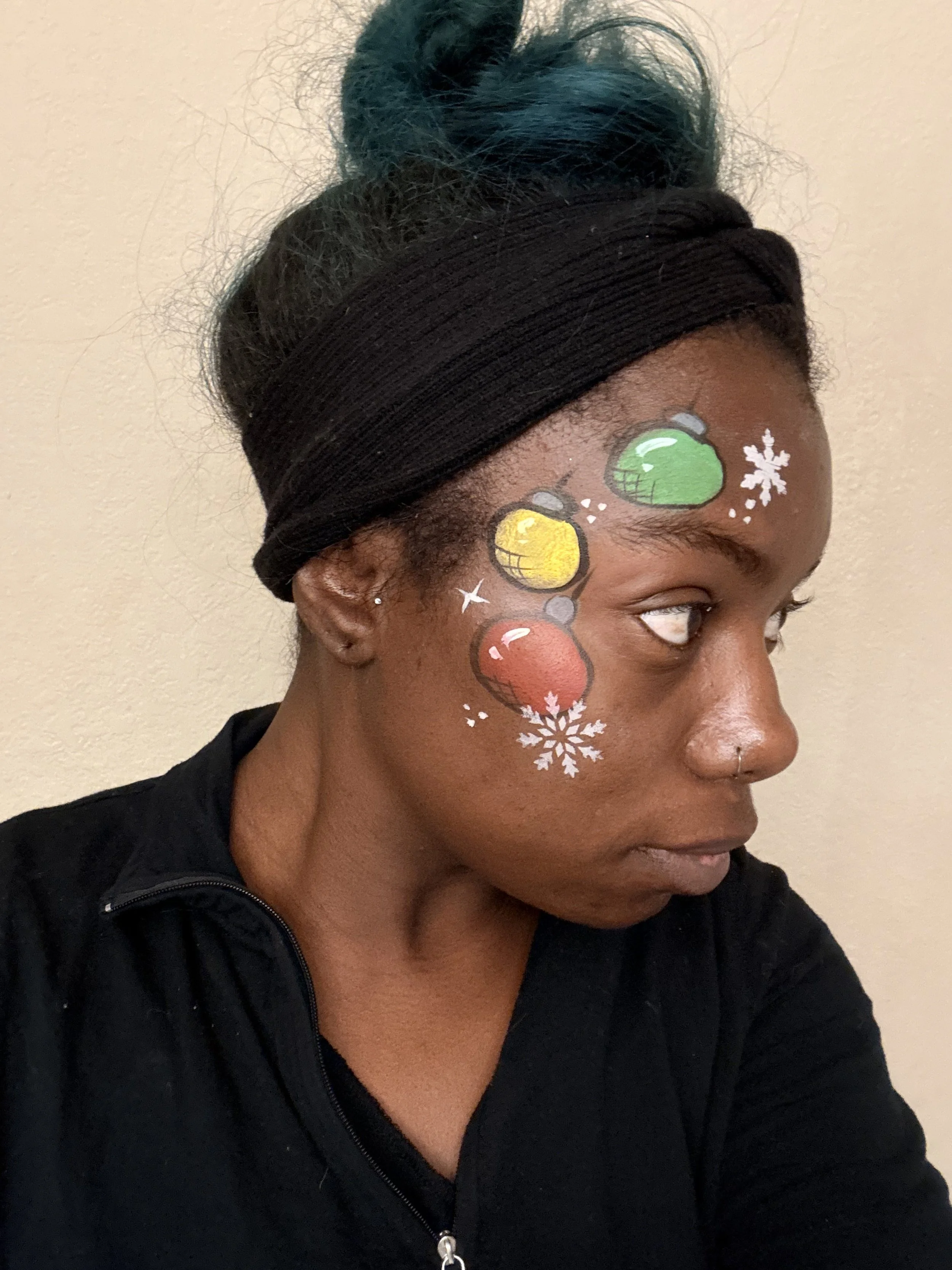 A woman with black hair tied in a bun, wearing a black headband and black zip-up top, has colorful Christmas ornaments and snowflakes painted on her face.