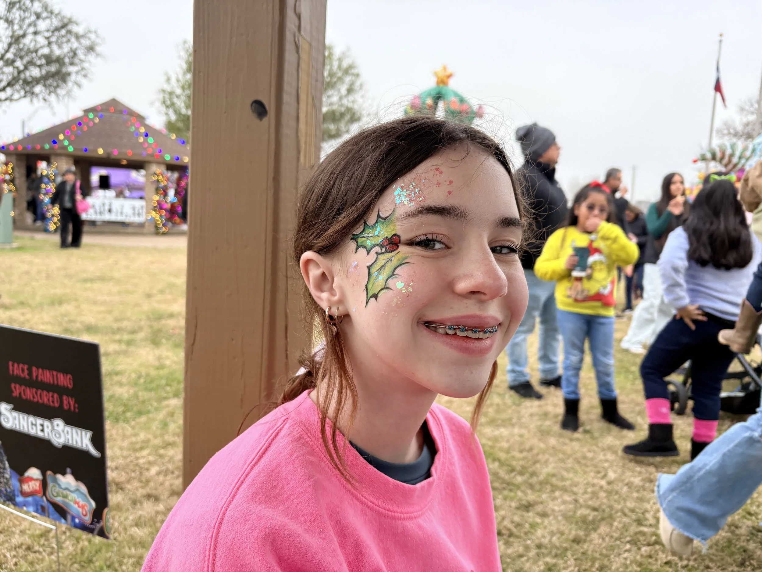 A girl with face paint of holly leaves and berries, smiling at a fair or festival with an outdoor stage decorated with Christmas lights in the background.