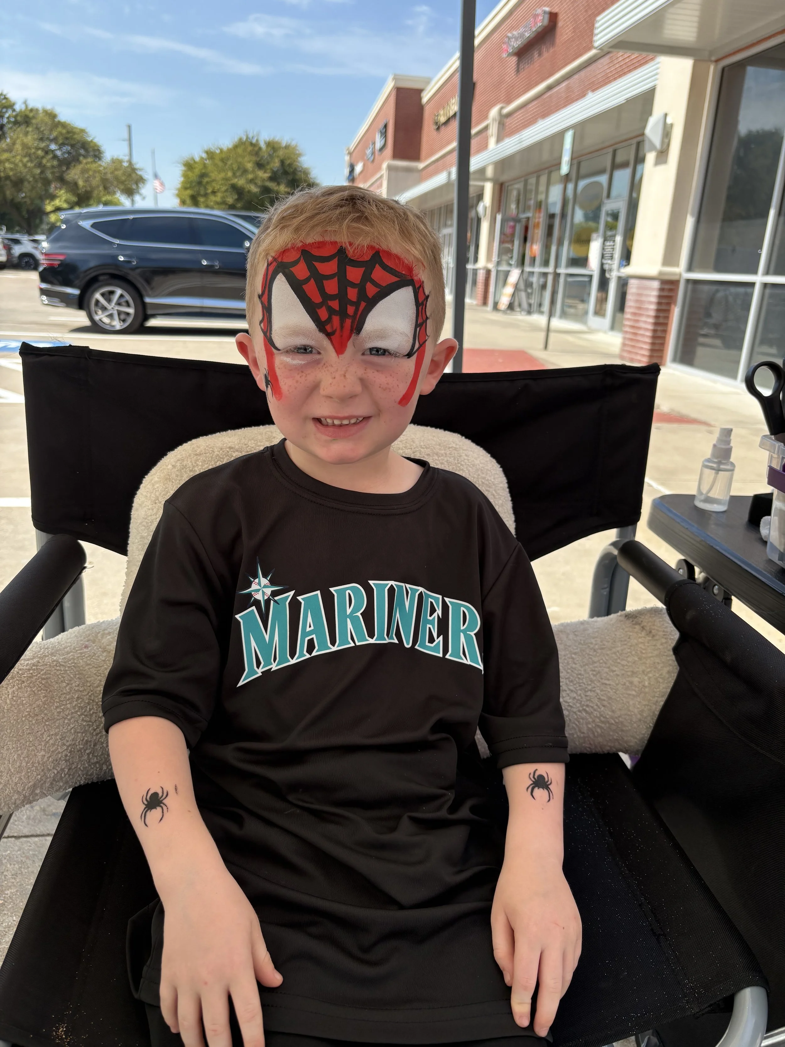 A young boy with face paint resembling Spider-Man, wearing a black Mariners shirt, sitting in a wheelchair outside a shopping center, smiling. He has temporary tattoos of spiders on his arms.