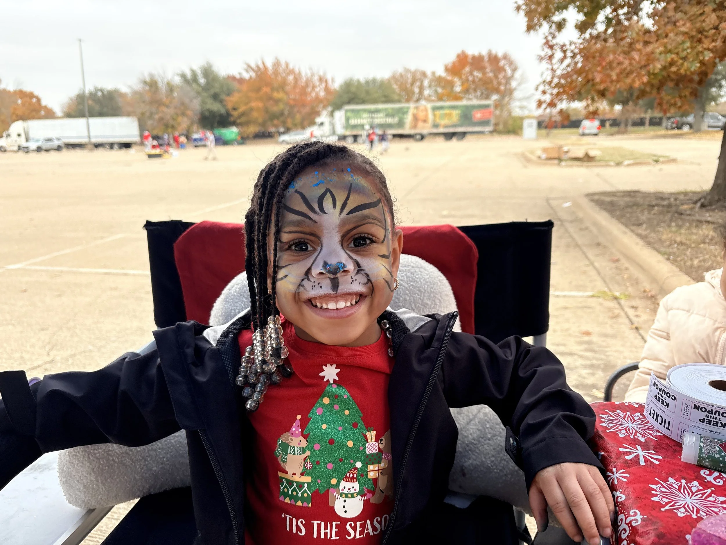 A young girl with face paint of a tiger, smiling, sitting at a table outdoors during the fall with trees showing autumn colors in the background.