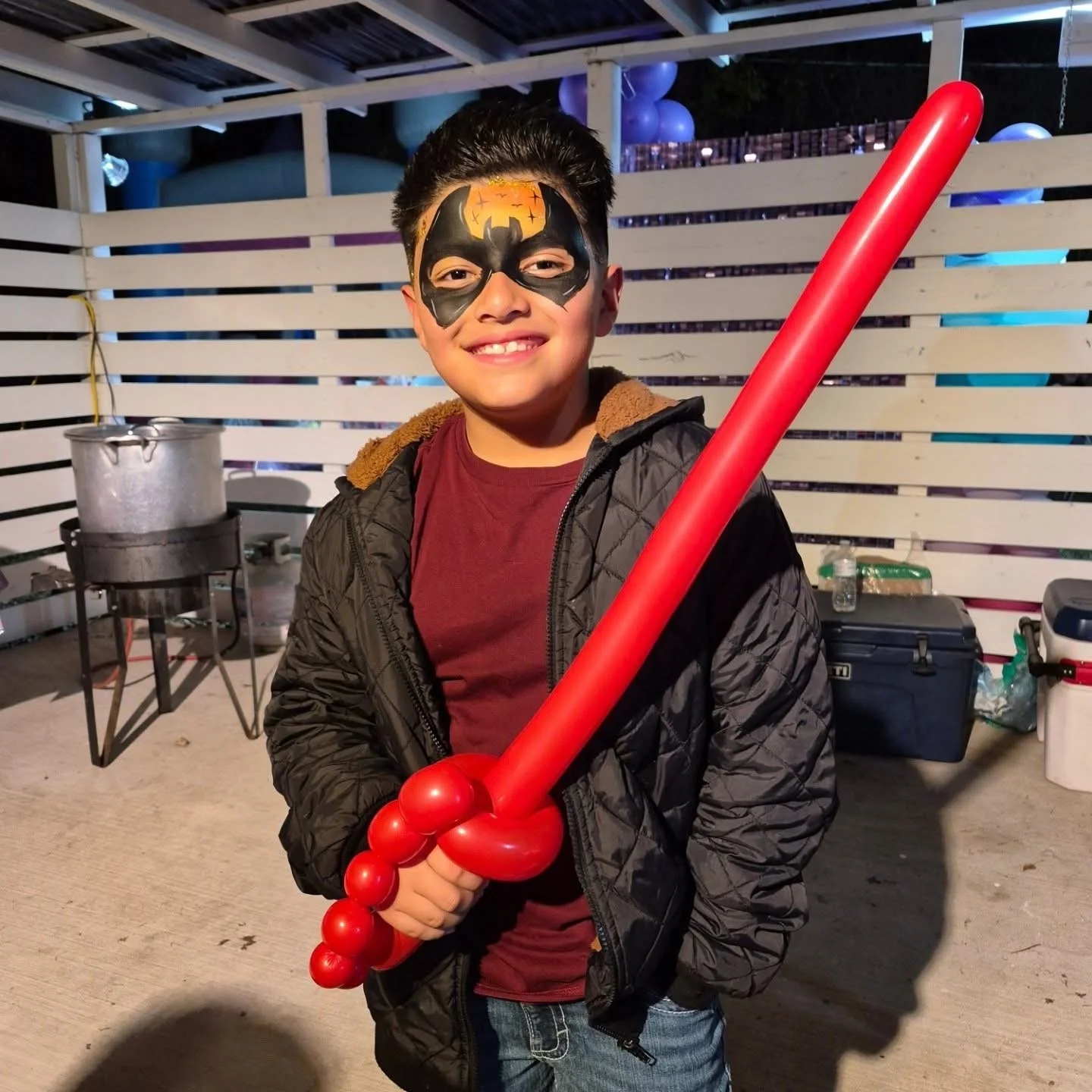A smiling boy with face paint resembling Batman, holding a red balloon sword, standing in a backyard with a white wooden fence, a large pot on a stand, and cooler boxes in the background.