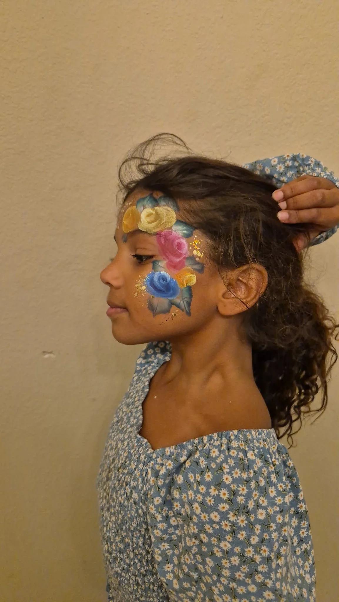 Girl with colorful floral face paint, wearing a blue dress with white daisies, standing against beige wall.