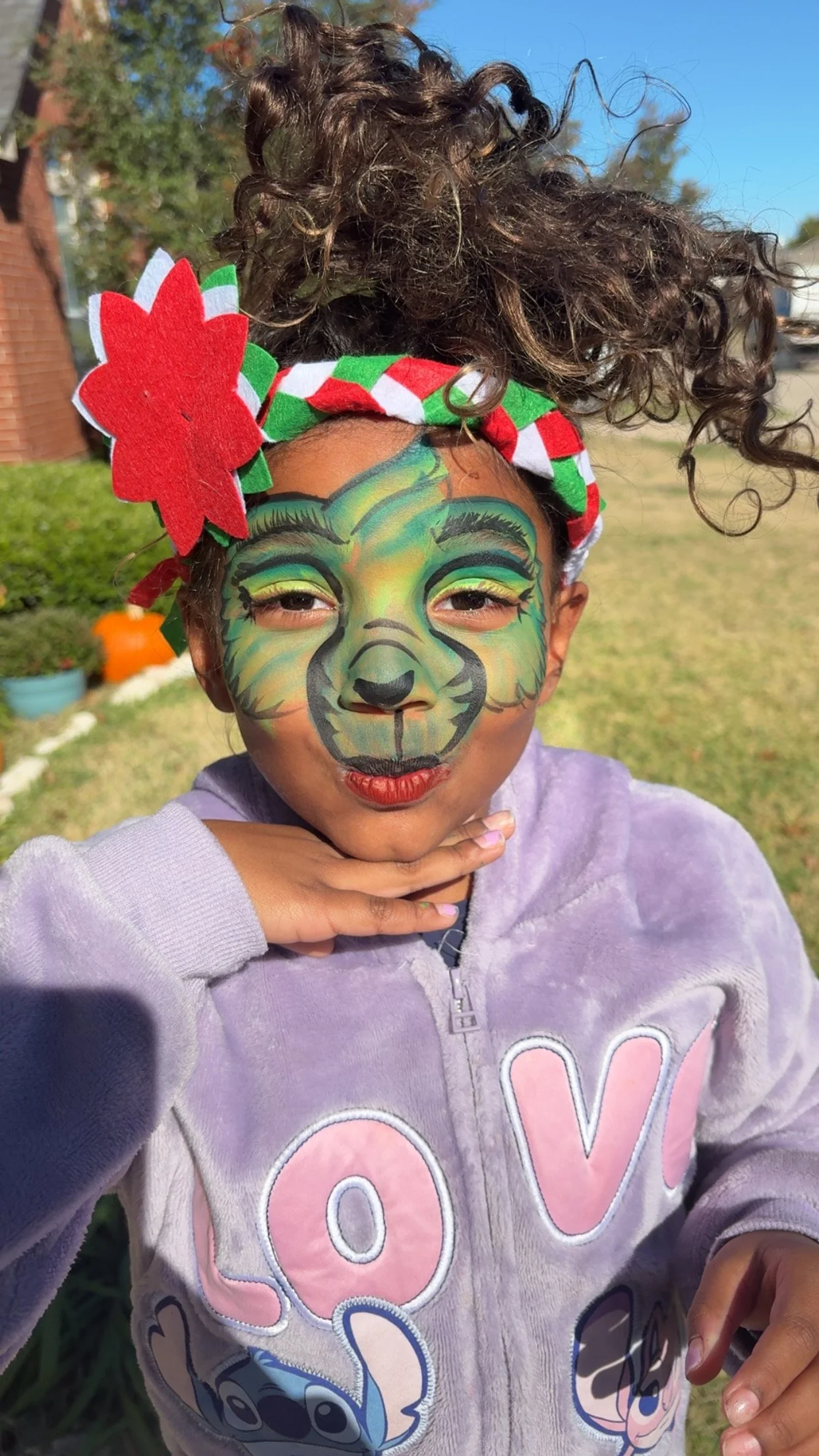 Child with face painting of a tiger, wearing a red, green, and white headband with a large red and green flower, outside on sunny day, wearing a purple hoodie with pink and blue lettering.