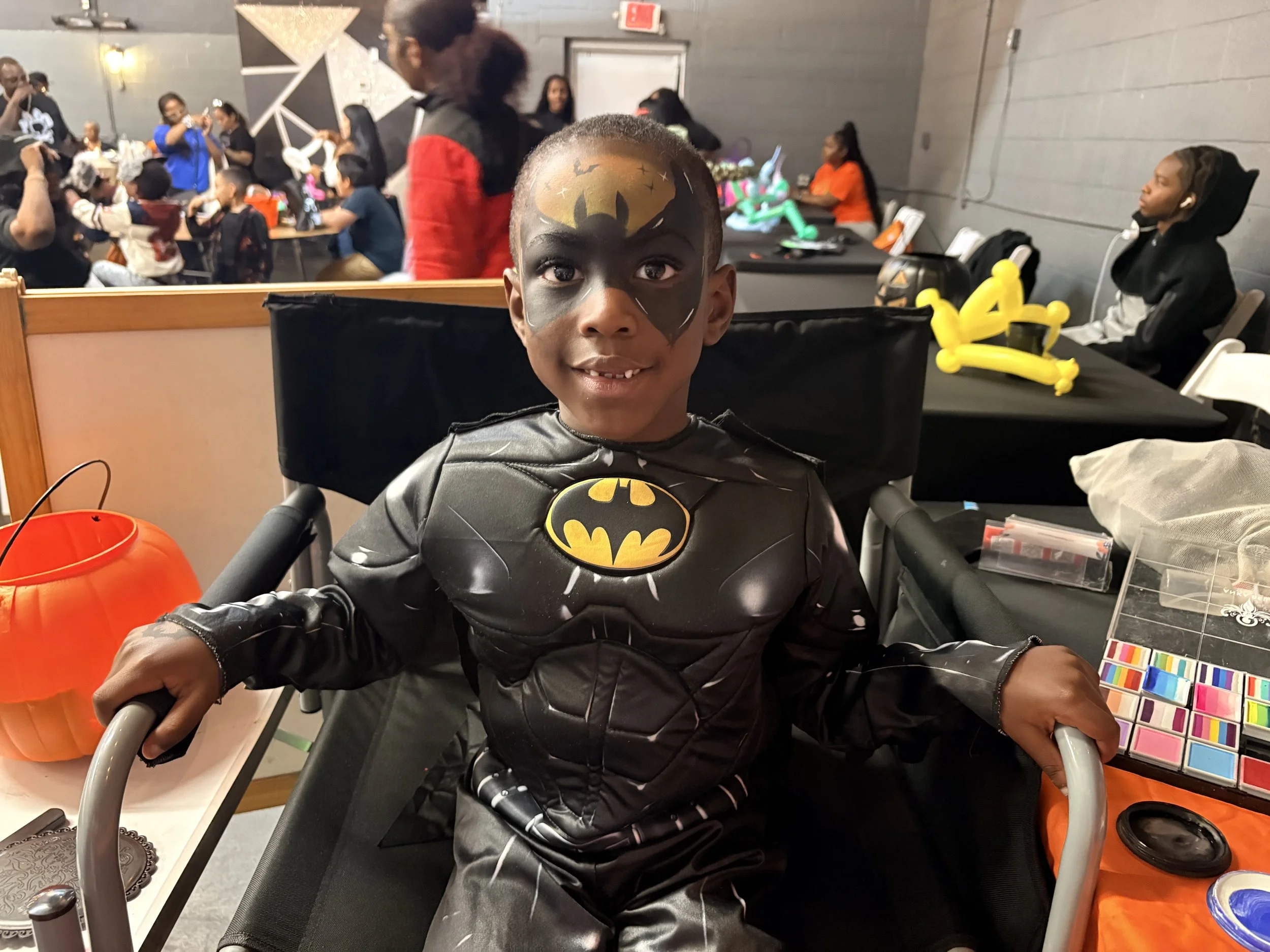 A young boy dressed as Batman with face paint, sitting at a table in a party or event setting, surrounded by decorations and other children and adults in the background.