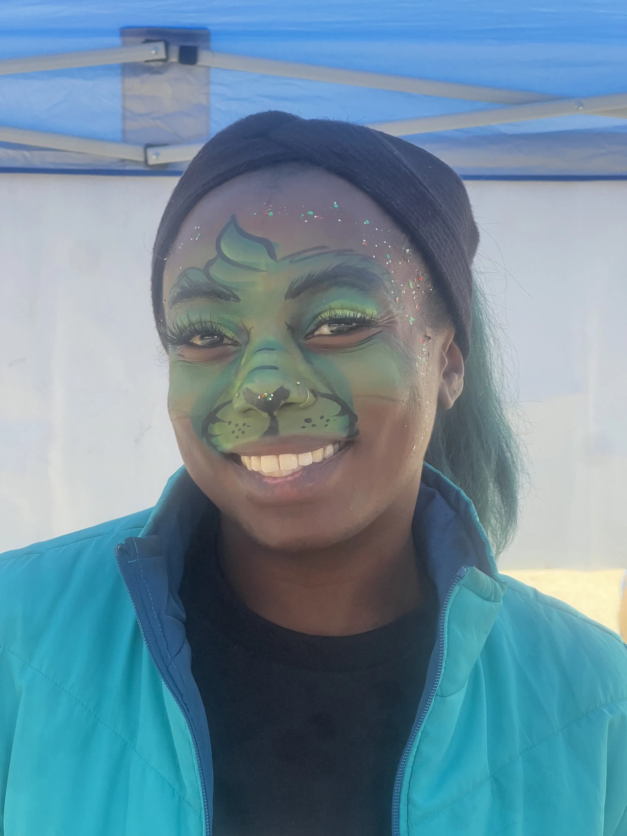 Person with face paint resembling a green and black animal, smiling, wearing a black headband and a blue jacket under a blue canopy.