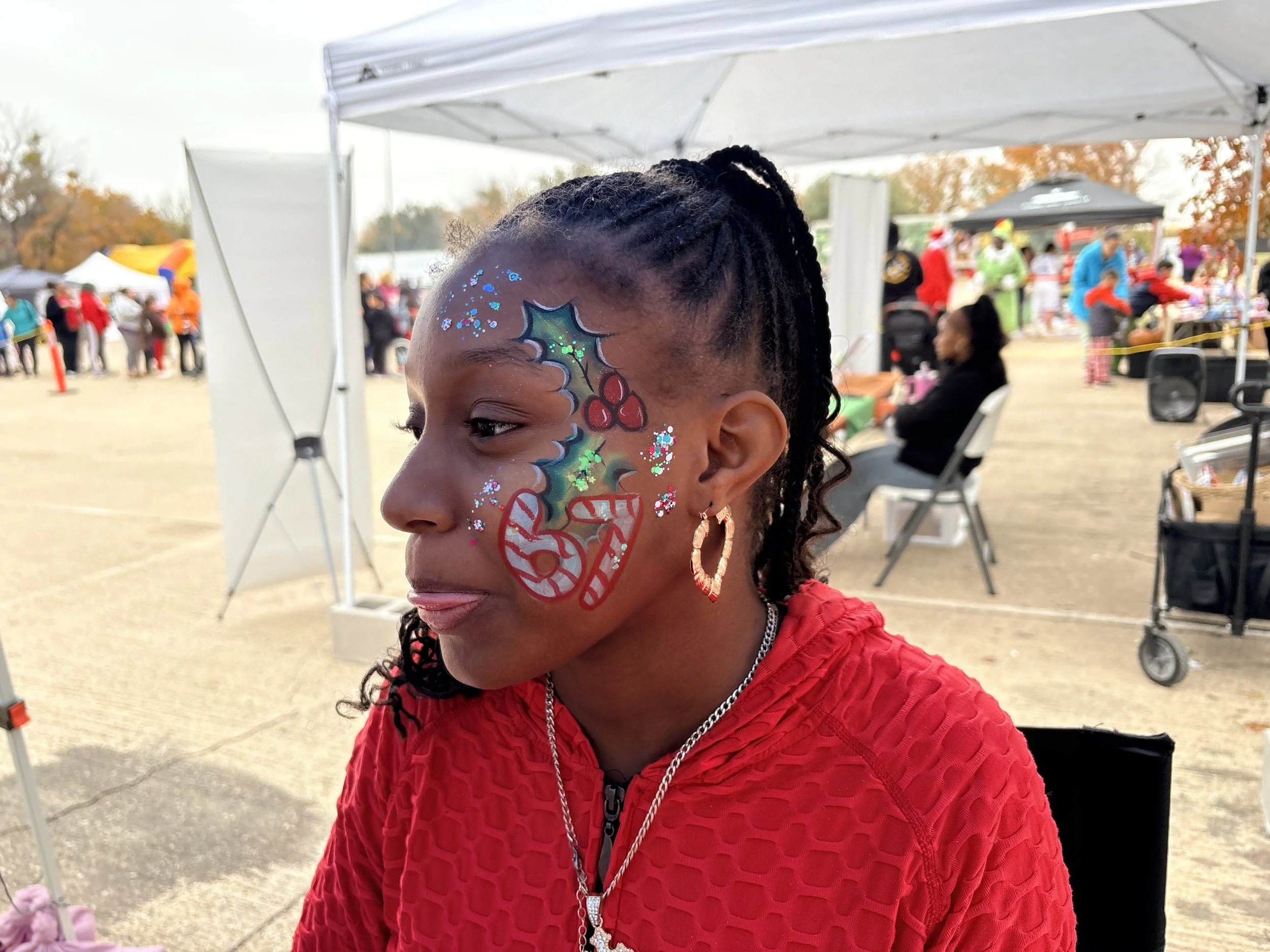 Young girl with Christmas face paint, including holly and candy cane designs, at an outdoor event with tents and people in the background.