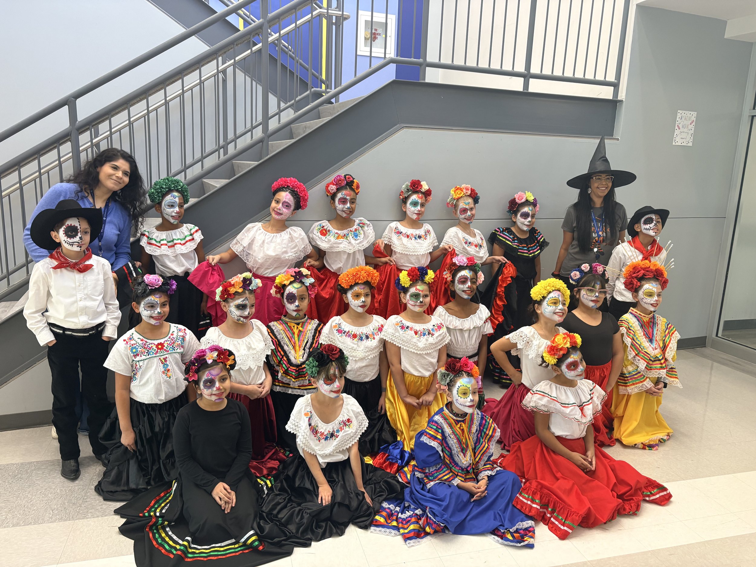Group of children and two adults dressed in traditional Mexican attire and face paint resembling sugar skulls, posing indoors near a staircase.
